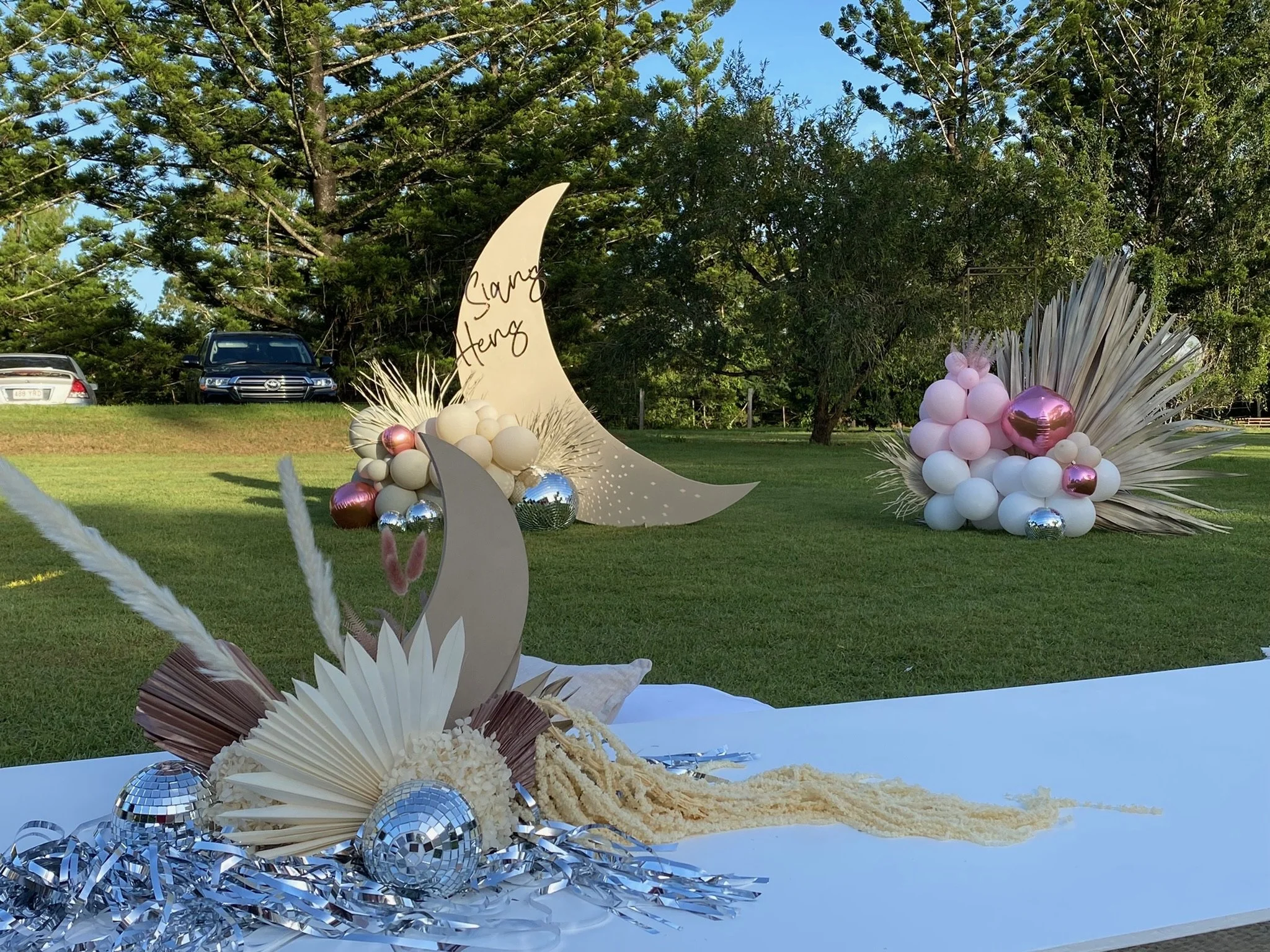 Decorative display featuring a large cream-colored crescent moon and a smaller cream-colored crescent moon with the names 'Siang' and 'Aeng' written on the larger moon. The display includes pink and white balloons, reflective silver and pink balls, a