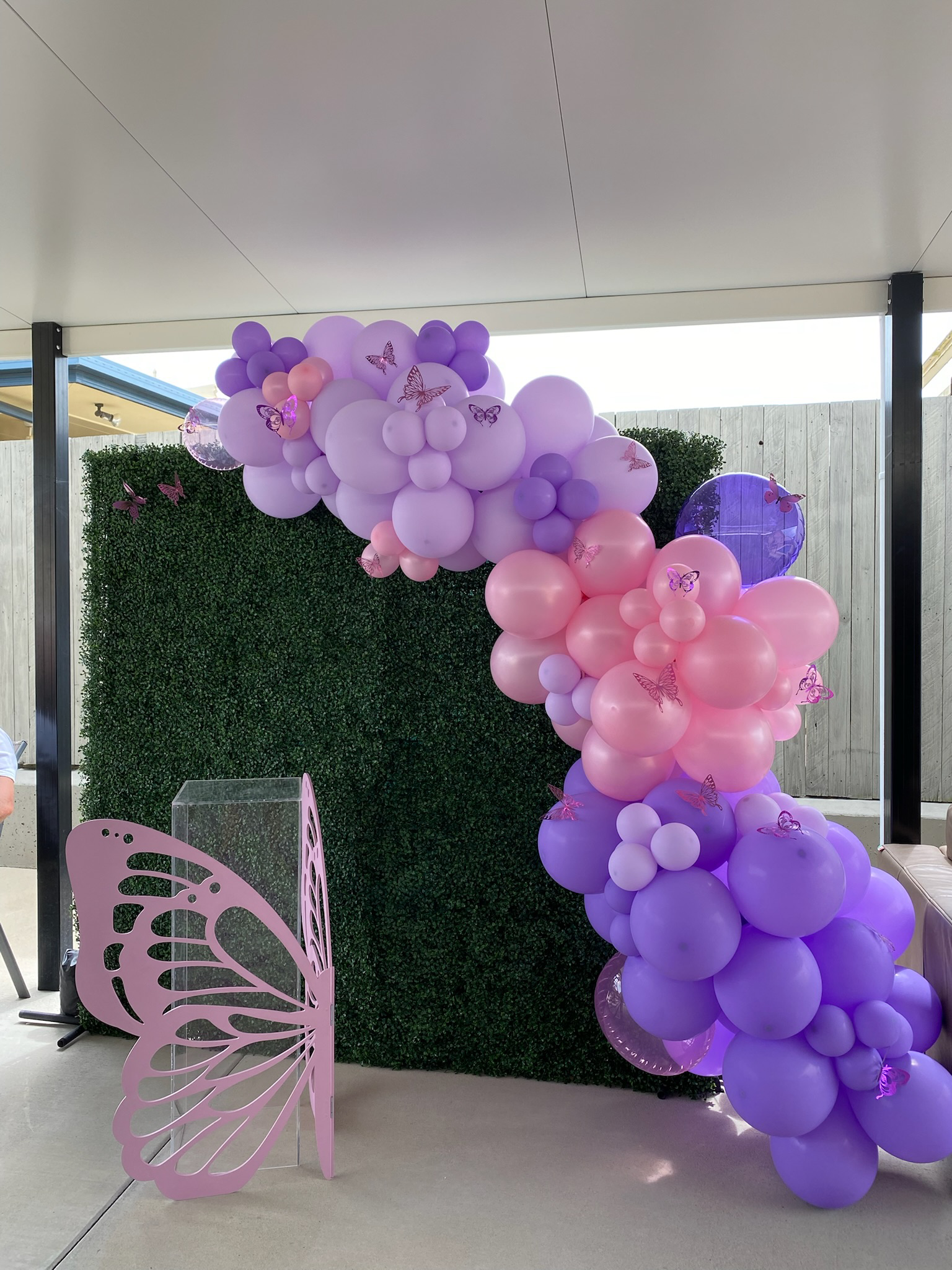 Decorative balloon arch with pink and purple balloons, decorated with pink and purple butterfly cutouts, in front of a green hedge backdrop.