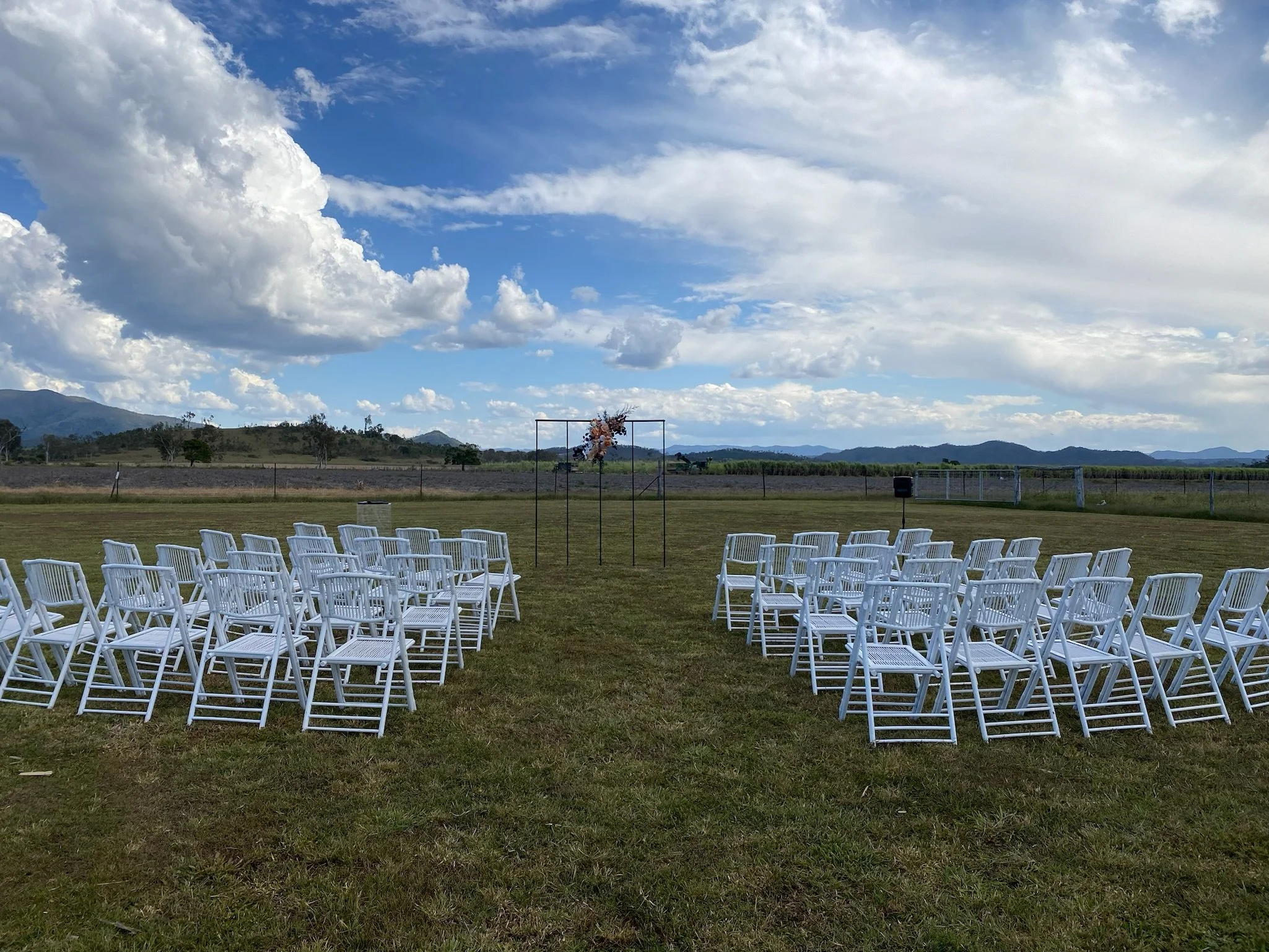 Outdoor wedding setup with white chairs arranged in two sections facing an arch decorated with pink flowers, on a grassy field under a partly cloudy sky with mountains in the distance.