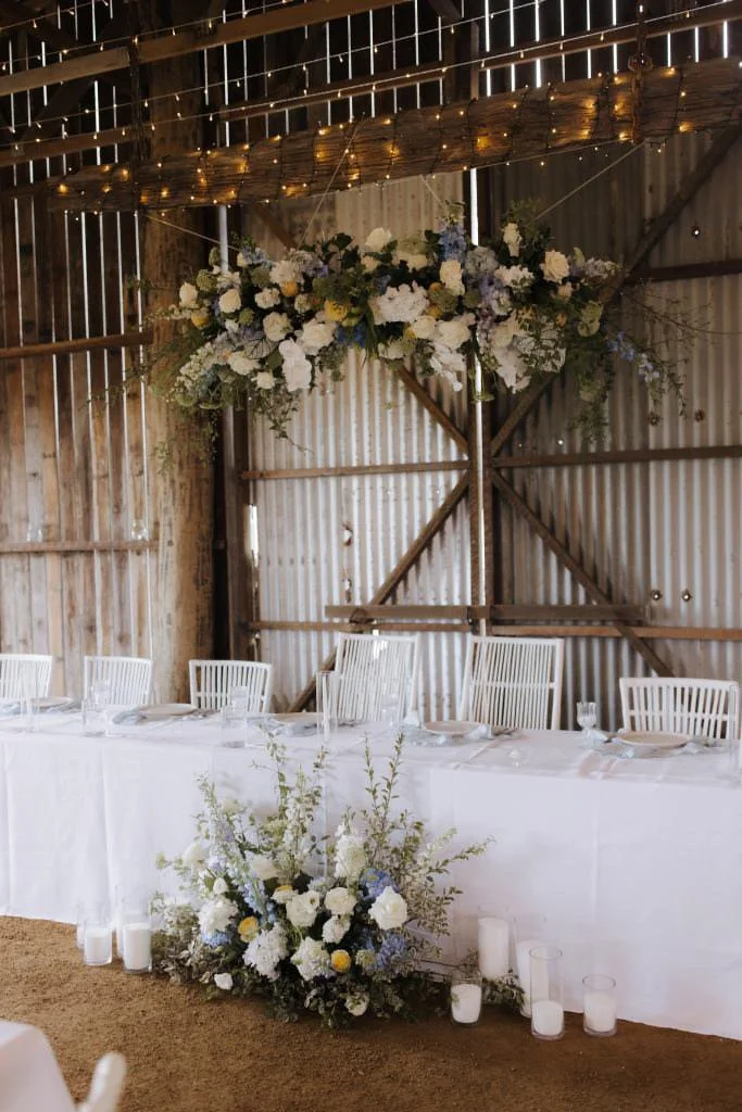 Decorated rustic barn wedding reception table with white tablecloth, floral centerpiece, hanging floral arrangement, candles, and white chairs.