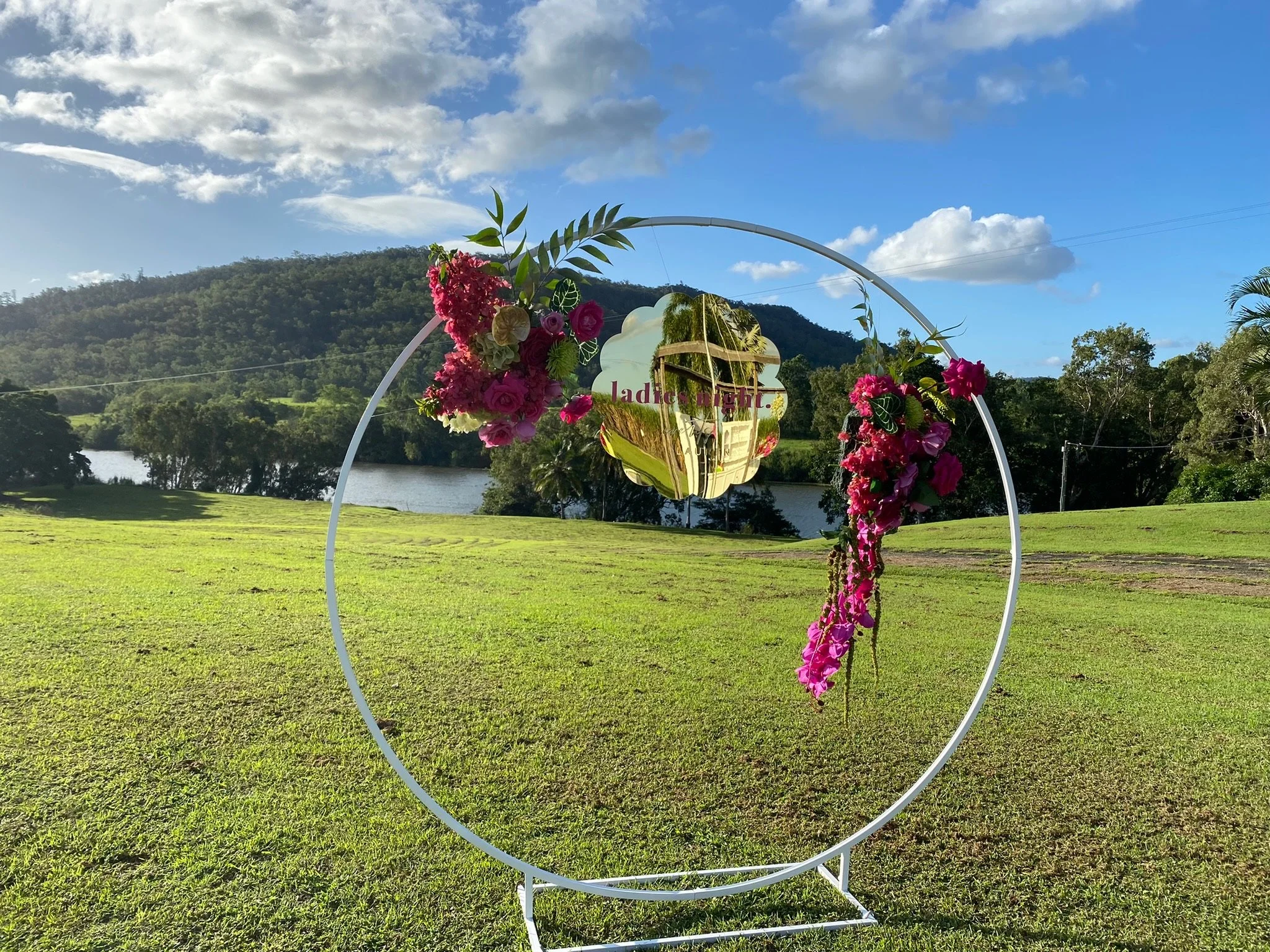 A decorative metal frame with pink and purple flowers and green leaves, set outdoors on a grassy field with a lake, trees, hills, and a partly cloudy sky in the background.