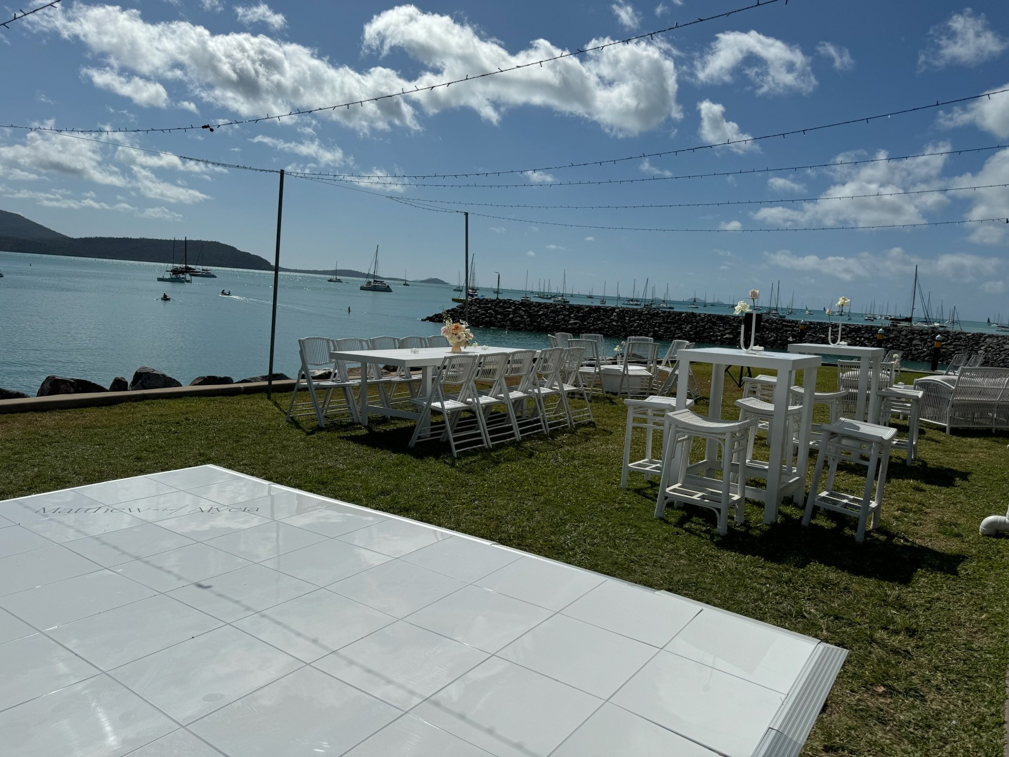 Outdoor event setup with white tables, chairs, and tall stools on grass by a waterbody, with sailboats and a breakwater in the background under a partly cloudy sky.