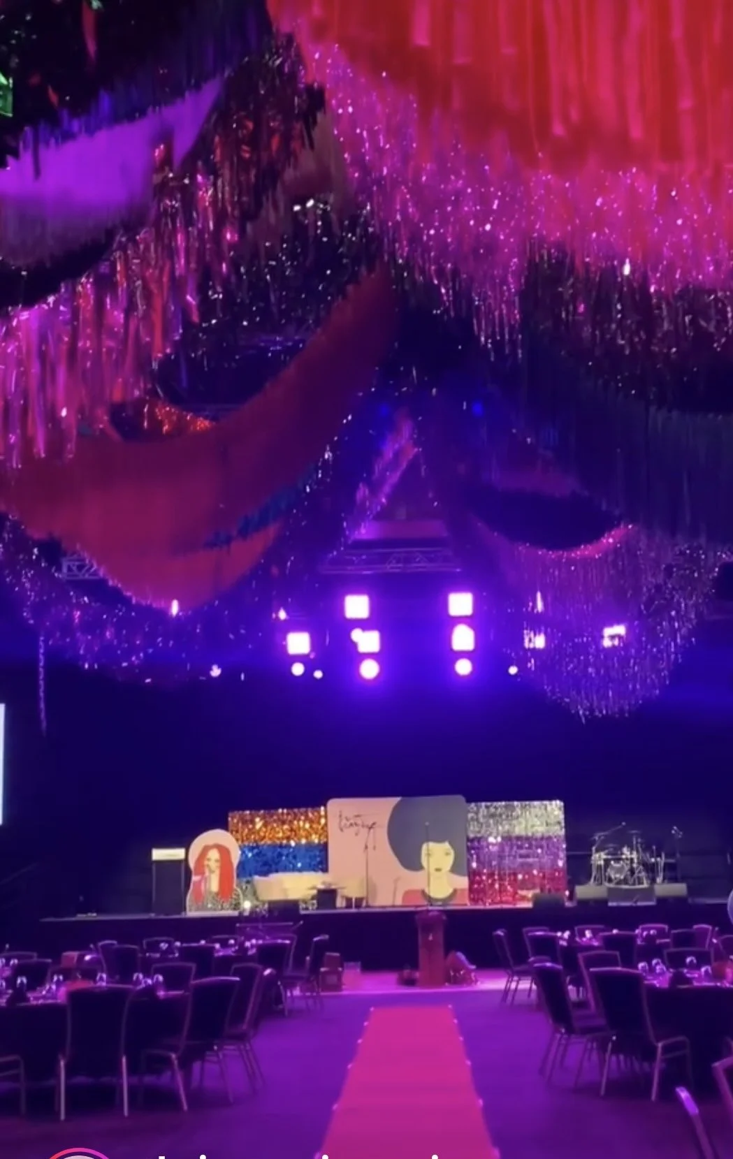 A decorated event venue with purple and pink lighting, hanging chandeliers, a stage with various artwork and decorations, and round tables with chairs set for an event.