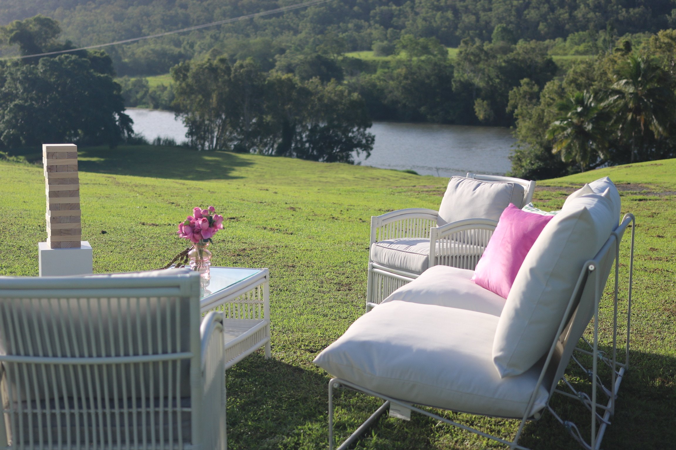 Outdoor patio with white wicker furniture, pink pillows, a glass table with a vase of pink flowers, overlooking a grassy landscape with trees and a river.