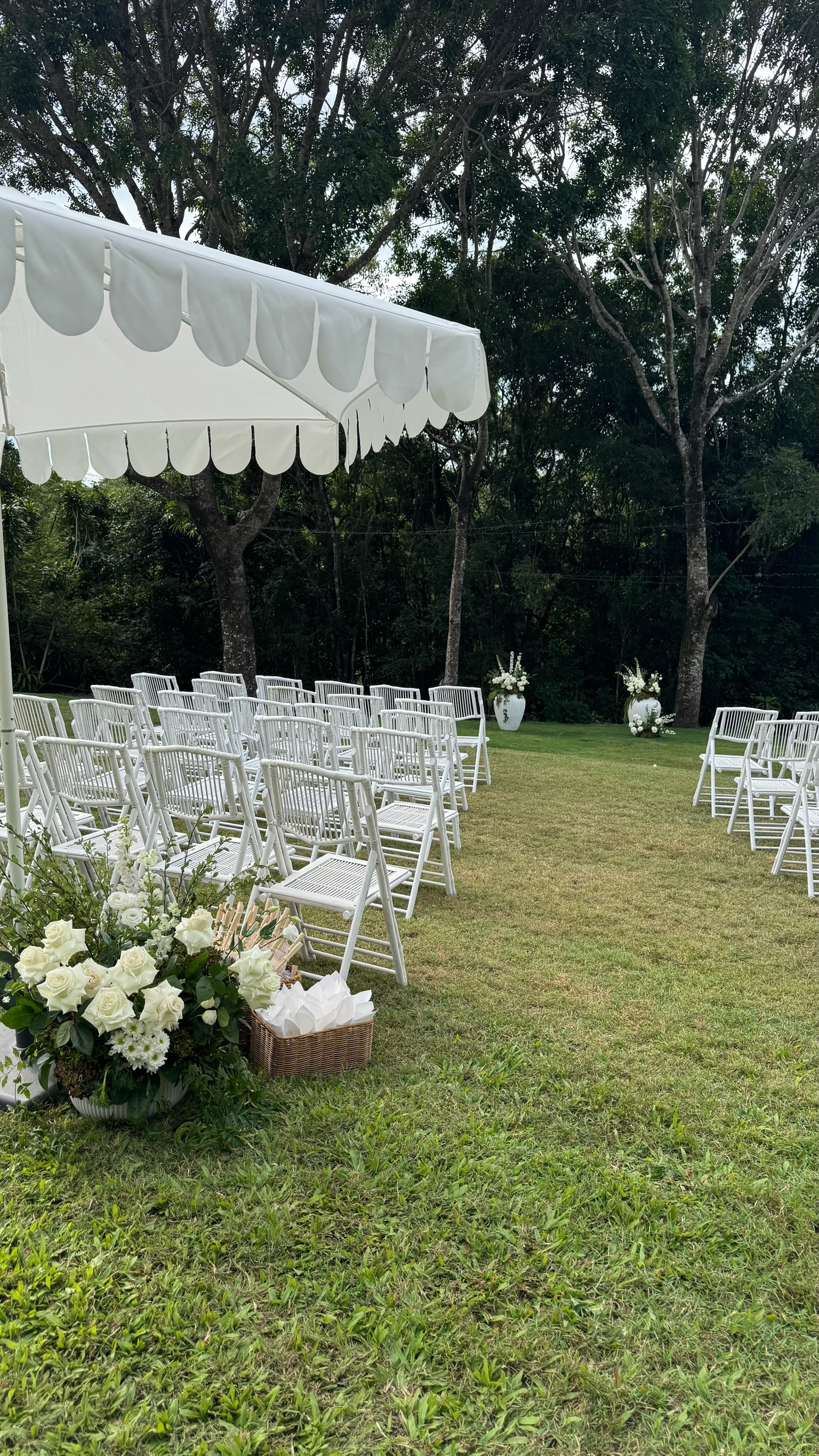Outdoor wedding setup with white chairs arranged on a grassy area under a white canopy, decorated with white flowers and large white vases with floral arrangements at the back, surrounded by trees.