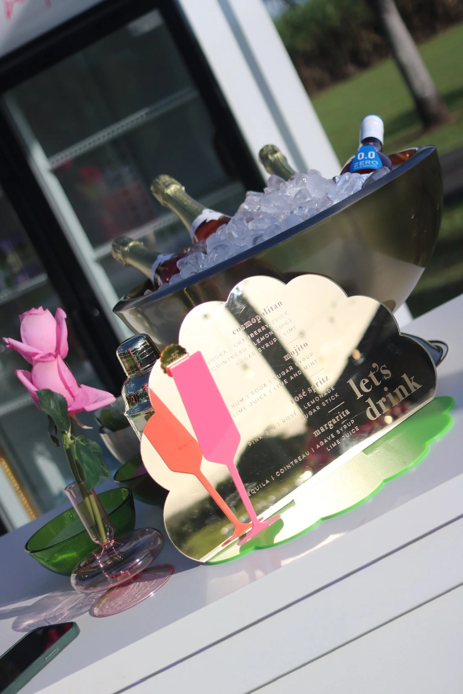 A metal bowl filled with ice and bottles of alcohol and small bottles of non-alcoholic beverages. To the left, there is a pink artificial flower in a small pink and green vase. The scene is outdoors with a building and trees in the background.