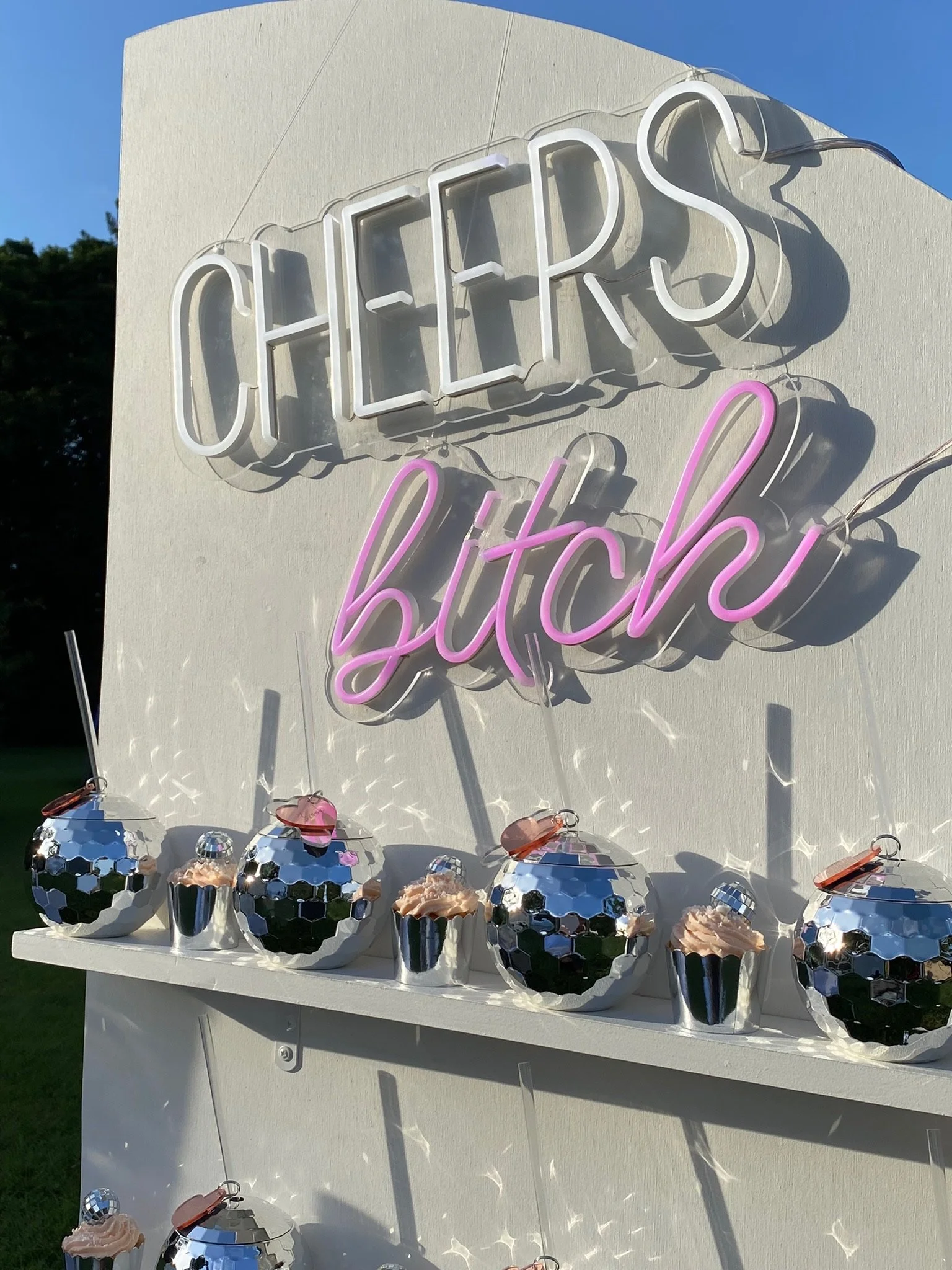 Neon sign reading 'CHEERS bitch' with mirrored disco ball cups filled with pink frosting lined up on a white stand.