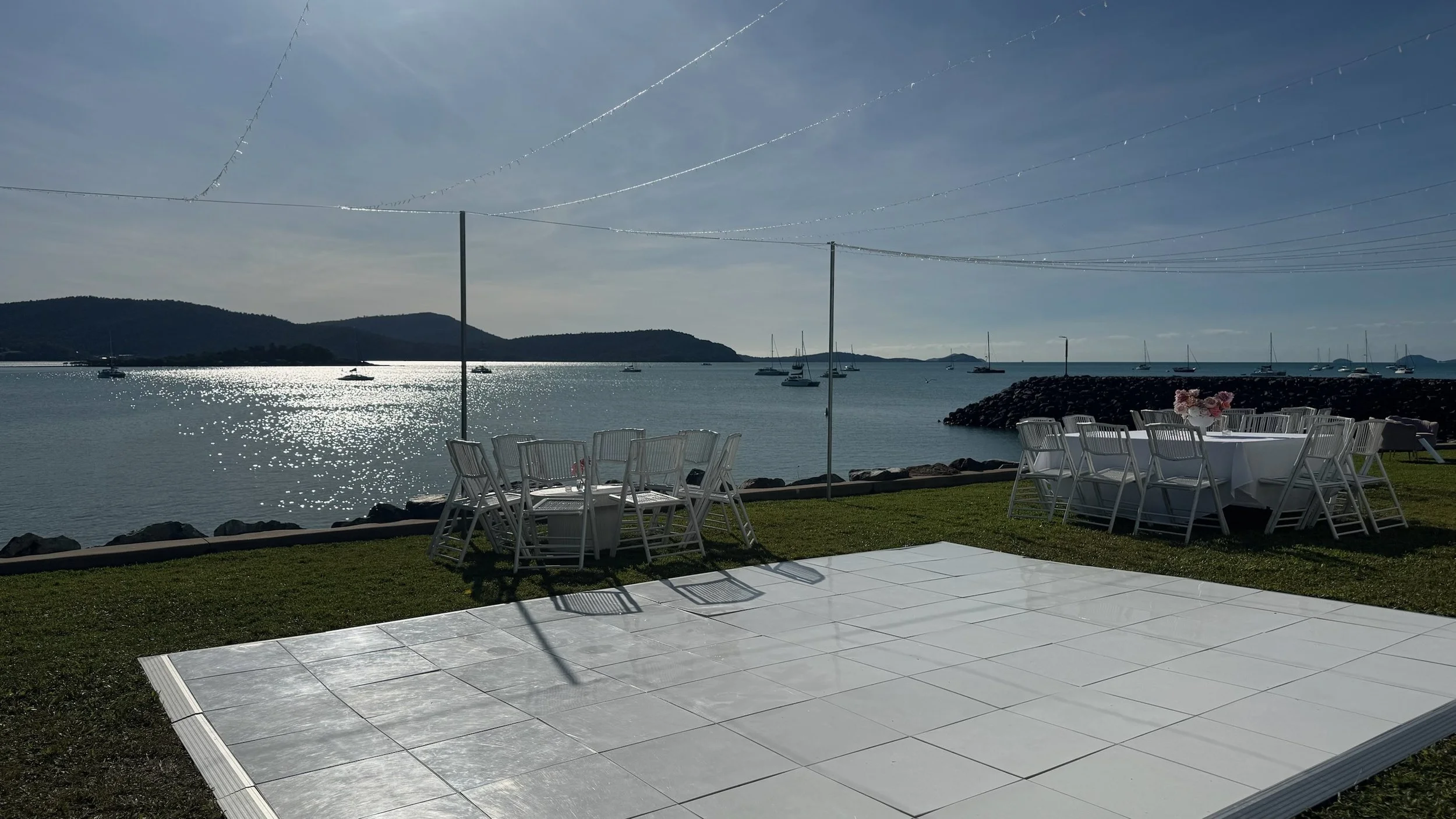 An outdoor event space near a body of water with white chairs and tables, some with pink flower arrangements, set on a grassy area and a white tile dance floor, with sailboats anchored in the water and mountains in the background.