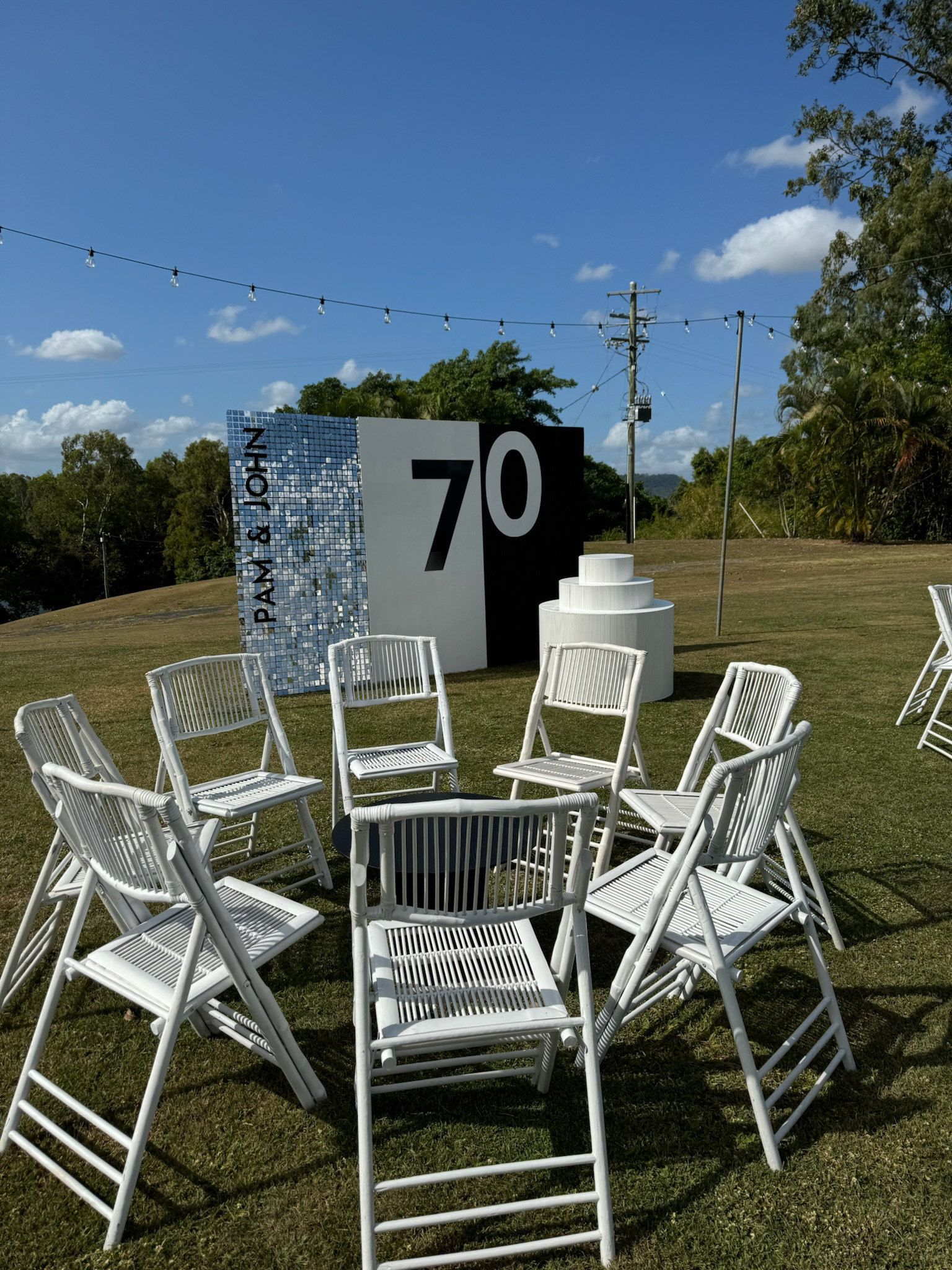 Outdoor event setup with white chairs arranged in a semi-circle on grass, a large sign displaying '70' and 'Pay & Join' behind, string lights overhead, and a backdrop of trees and a blue sky with clouds.