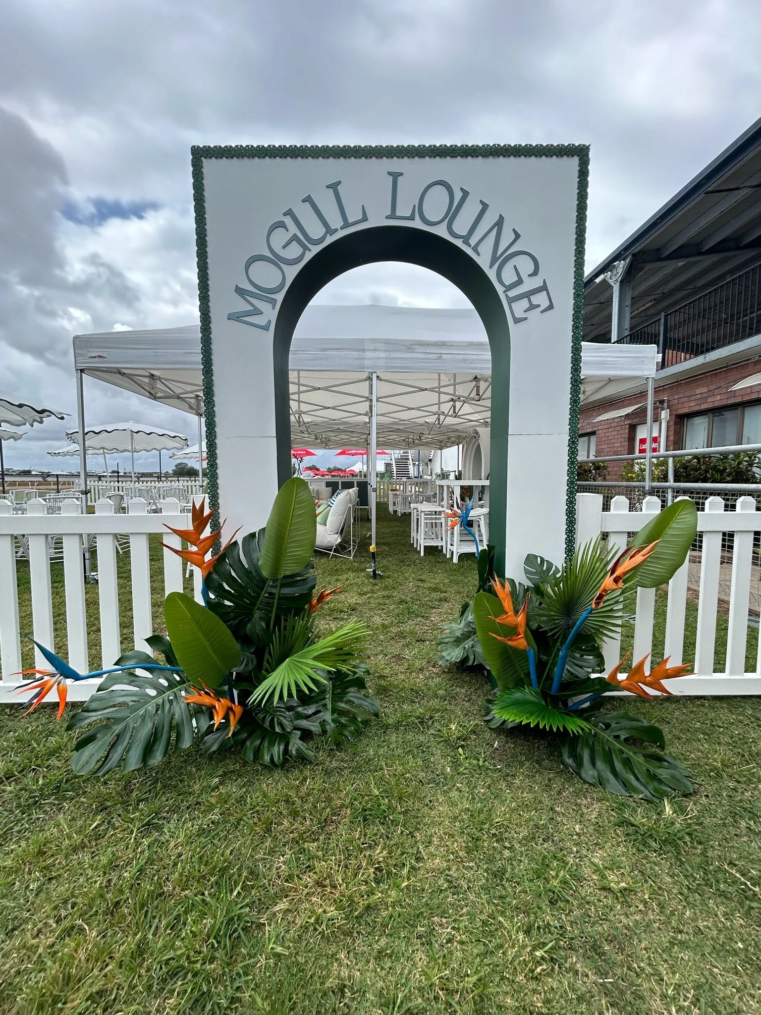 Entrance archway labeled 'Mocalull Lounge' with tropical plants and flowers in front, and outdoor seating area with tables and umbrellas in the background.