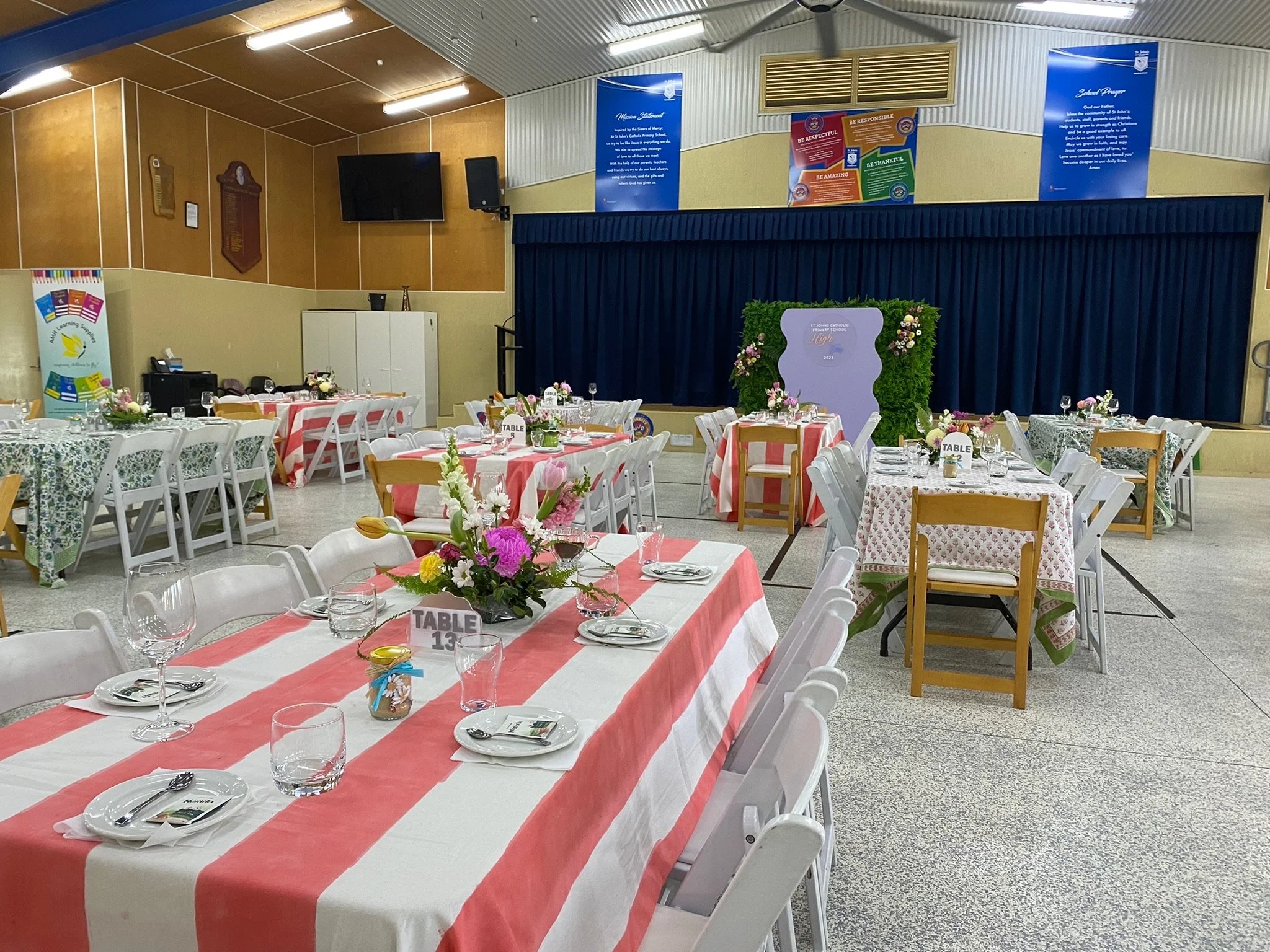 Decorated banquet hall with round and rectangular tables covered with floral and striped tablecloths, set with glassware, napkins, plates, and floral centerpieces, prepared for a formal event.