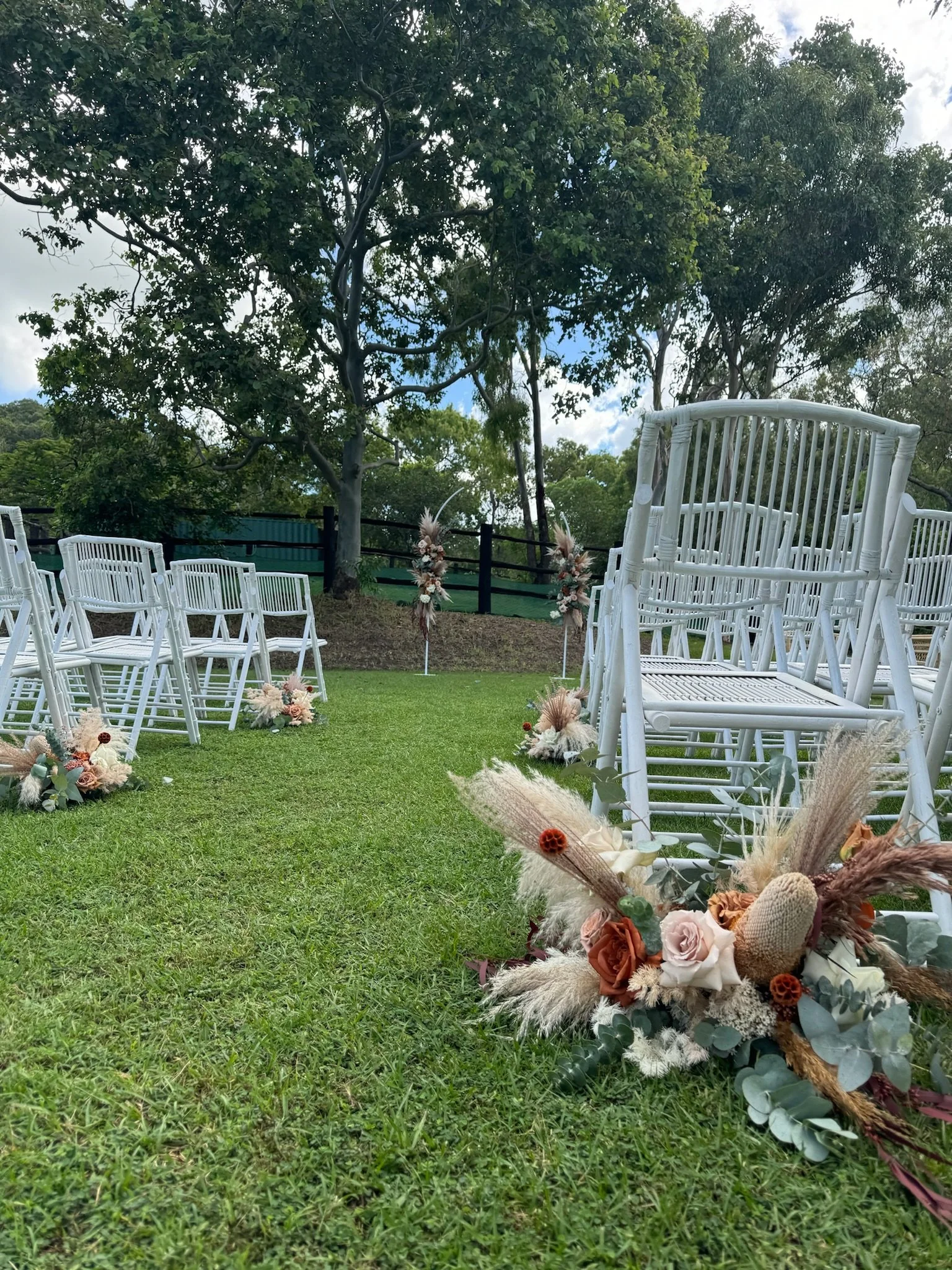 Outdoor wedding ceremony setup with white chairs arranged on green grass, decorated with floral arrangements and pampas grass, in a natural setting with trees and cloudy sky in the background.