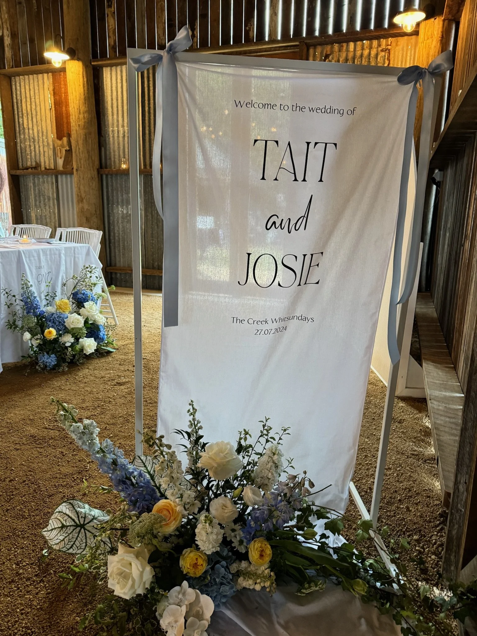 Wedding sign welcoming guests to the wedding of Tait and Josie, with floral arrangements at the base, inside a rustic venue with wood and corrugated metal walls.