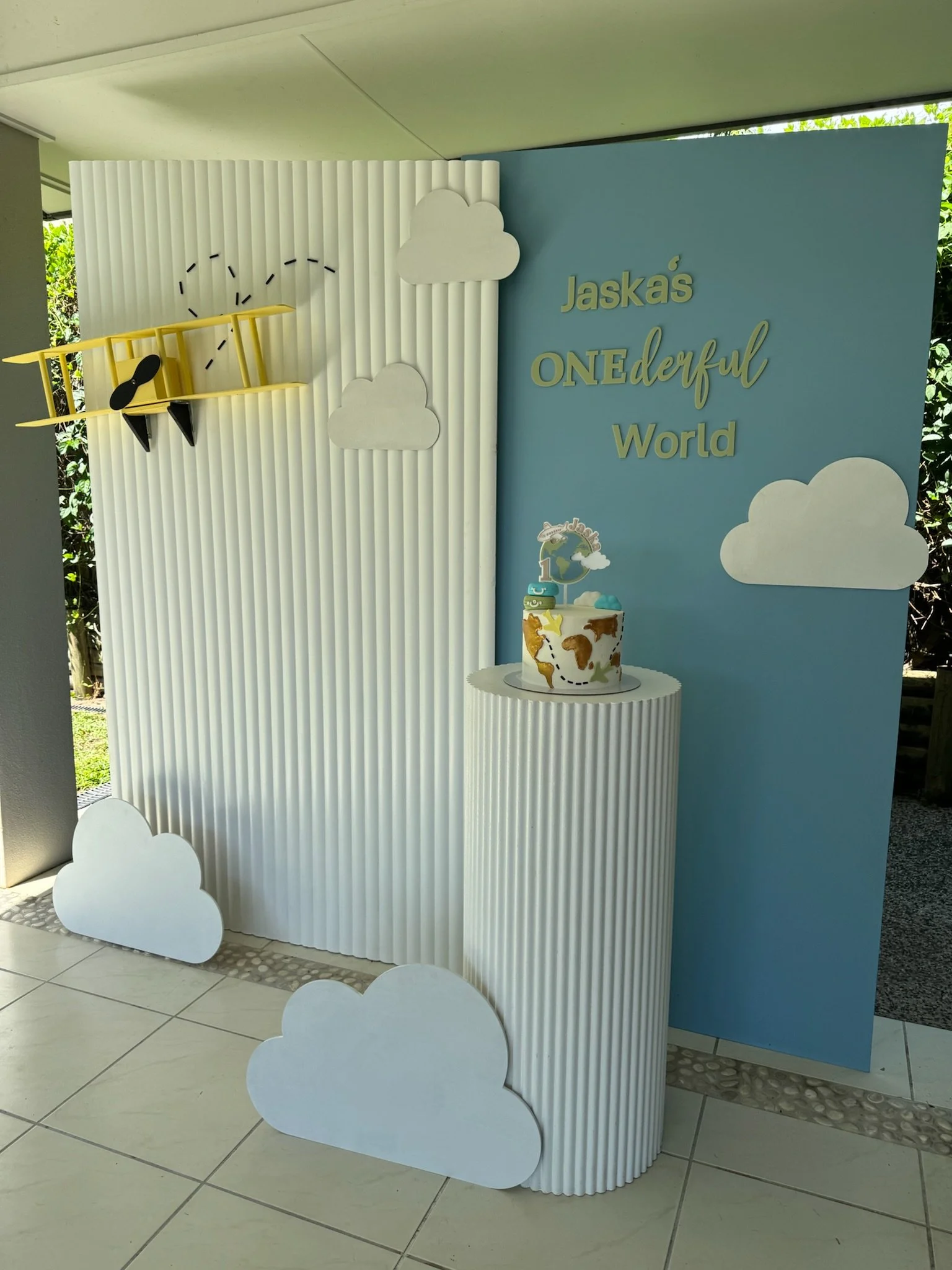 Decorative cloud-themed backdrop with a cake, a yellow toy airplane, and a sign reading "Jaskás ONEderful World."