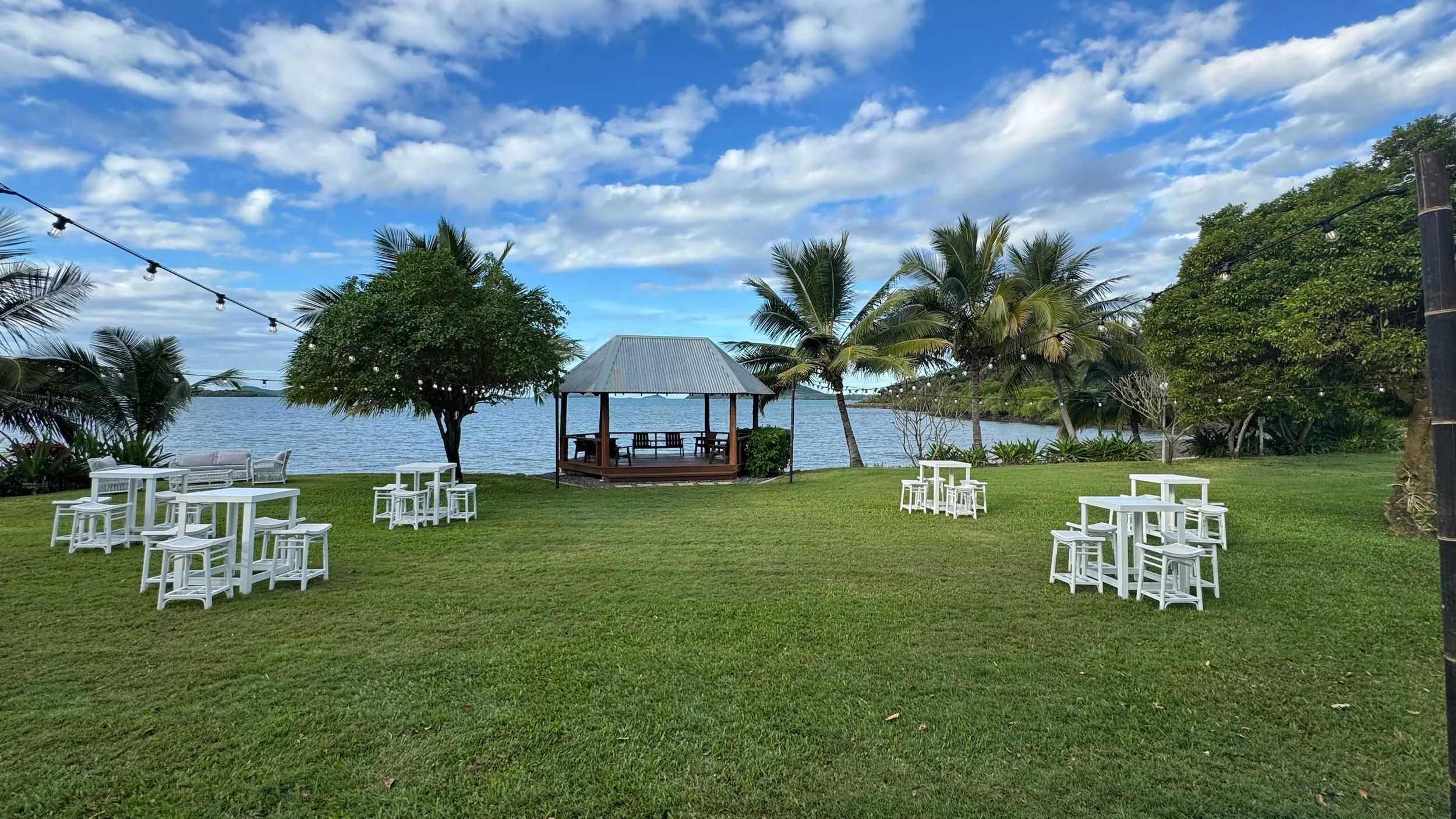 A scenic outdoor event space near a body of water with green grass, several white tables and stools, a wooden gazebo with a metal roof, string lights, trees, and palm trees under a partly cloudy sky.