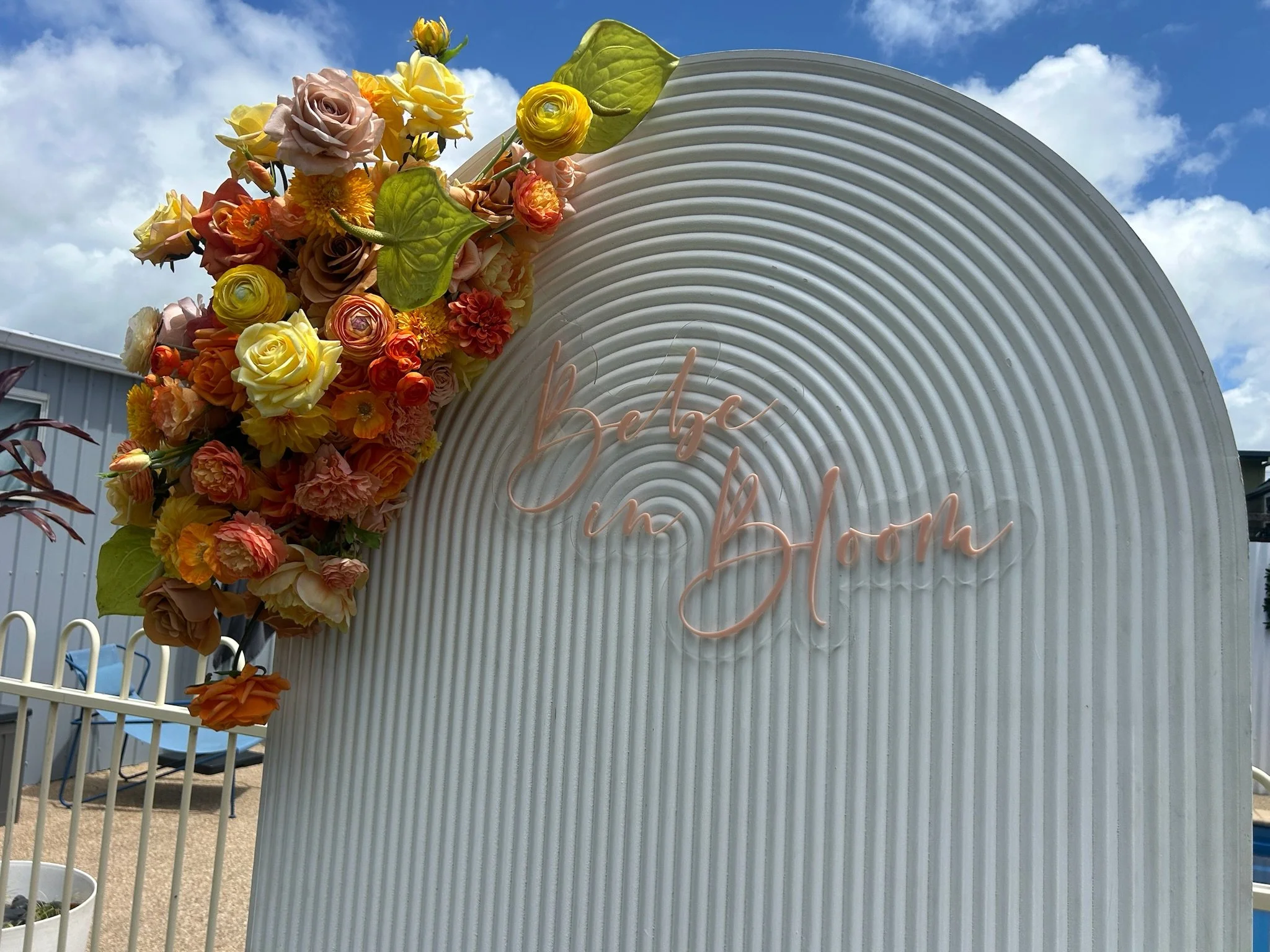 Outdoor decorative sign with a floral arrangement reading "Baby in Bloom" against a background of blue sky with clouds.