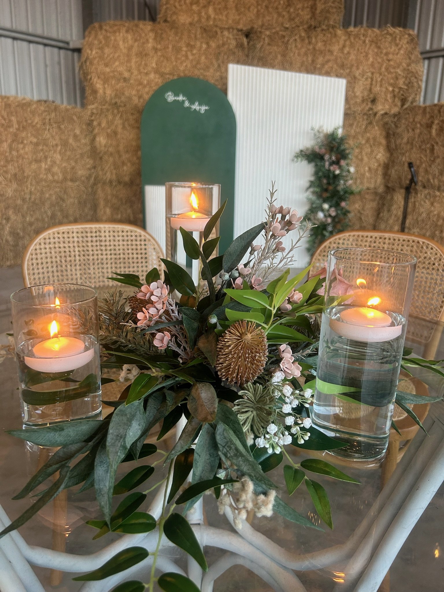 A floral centerpiece with candles on a glass table, with hay bales and decorative panels in the background.