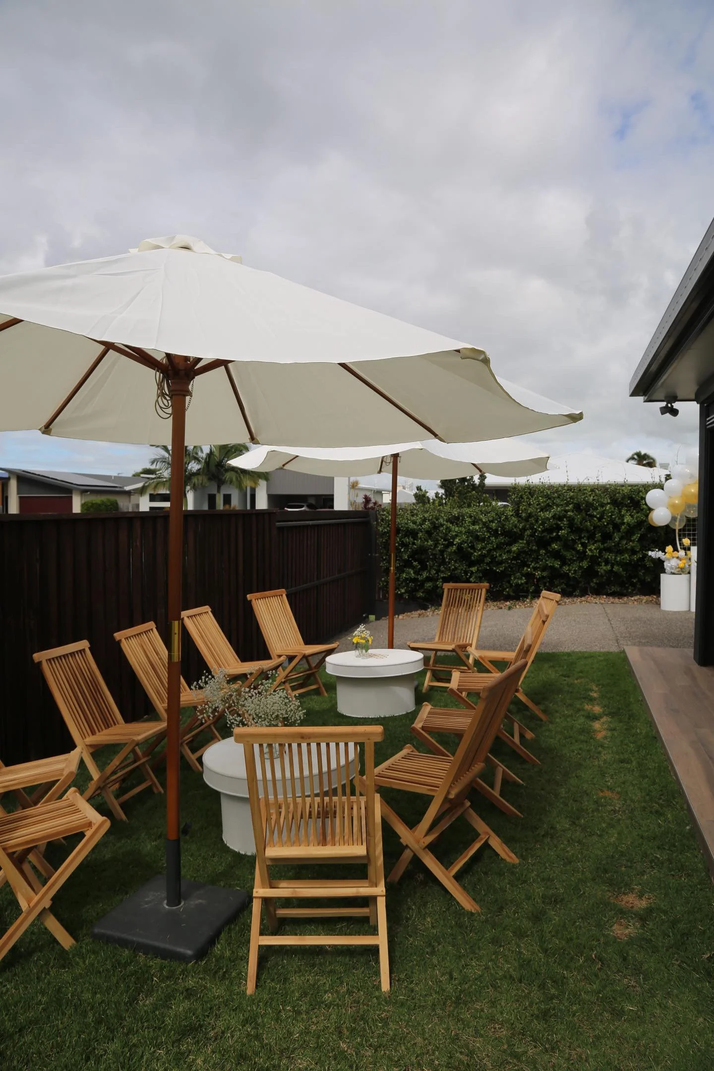 Outdoor patio area with wooden chairs and white tables under large white umbrellas, surrounded by a wooden fence and greenery, with cloudy sky overhead.