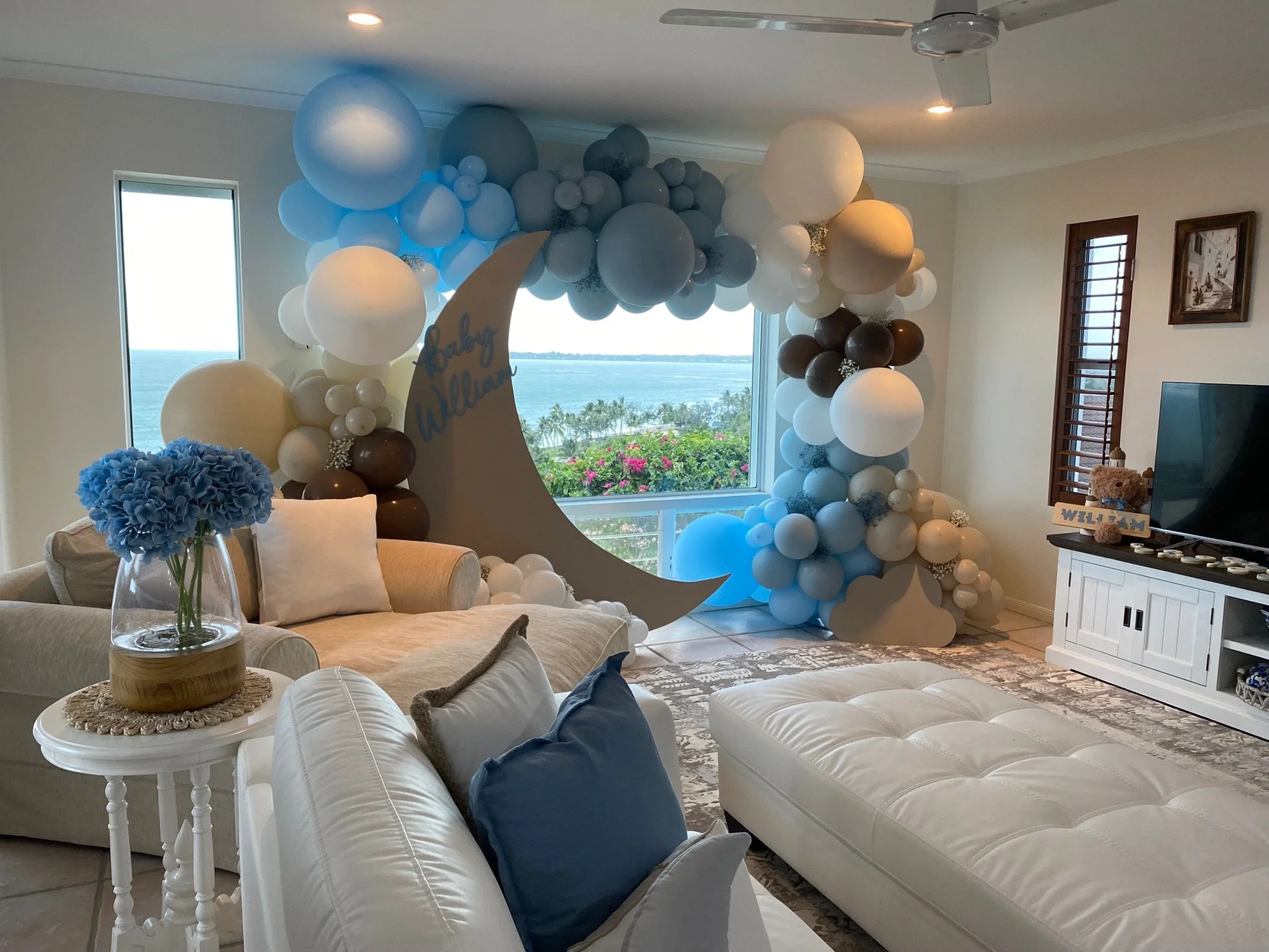 Living room decorated with a balloon arch in shades of blue, white, and brown, featuring a crescent moon cutout with the words 'baby William.' The room has large windows with a view of the ocean, a white ottoman, a beige sofa with blue and beige pill