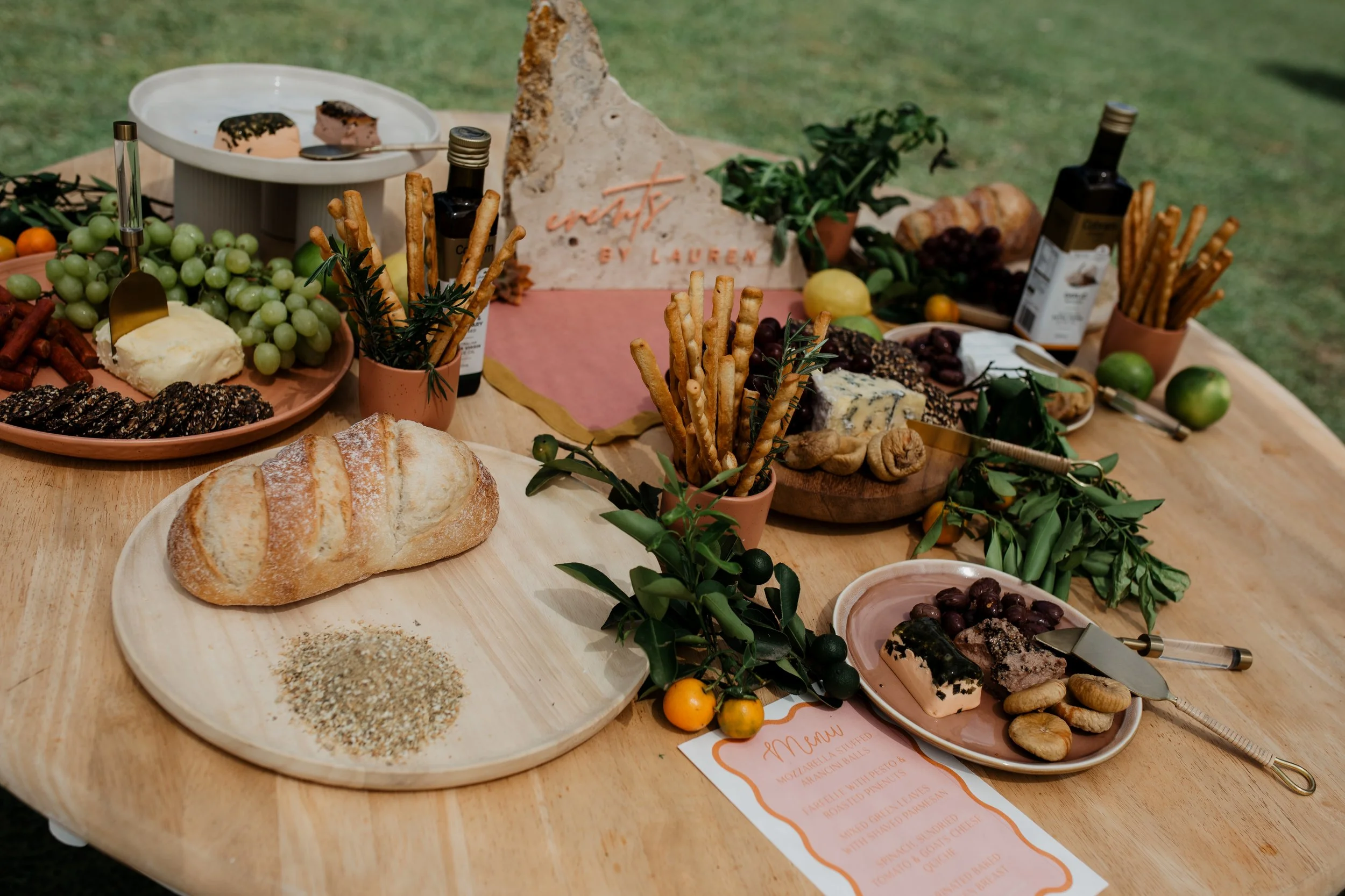 An outdoor food spread featuring bread, cheese, grapes, crackers, and various snacks on a wooden table decorated with greenery, citrus fruits, and a pink menu card.