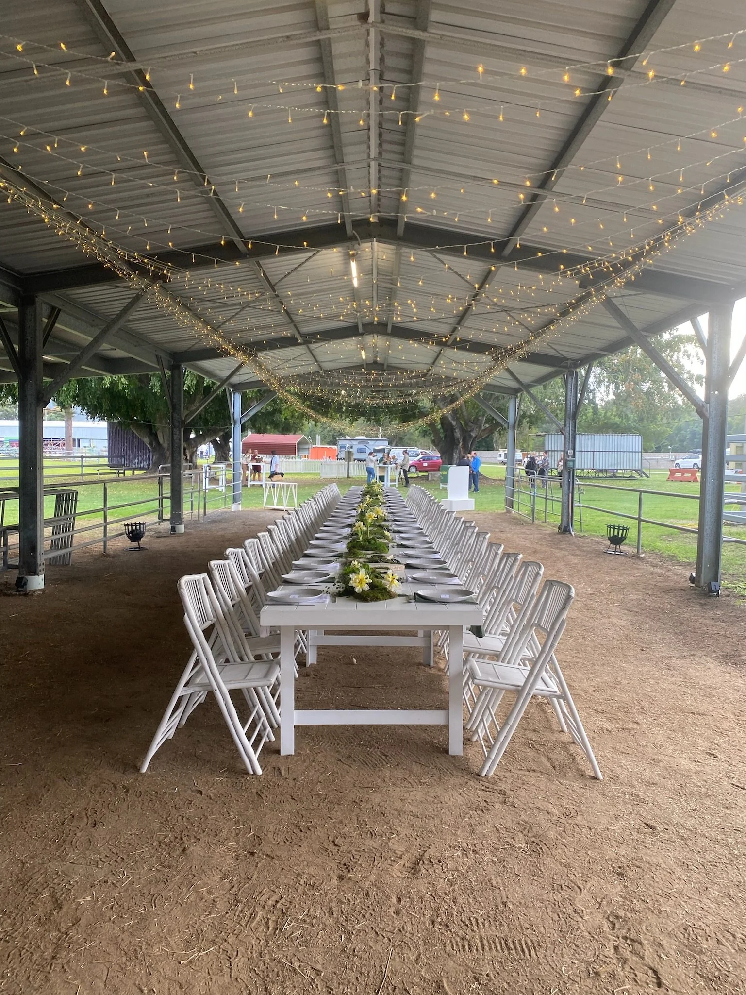 Long rectangular white table set for a formal event with white chairs, floral centerpieces, under a decorated outdoor pavilion with string lights.