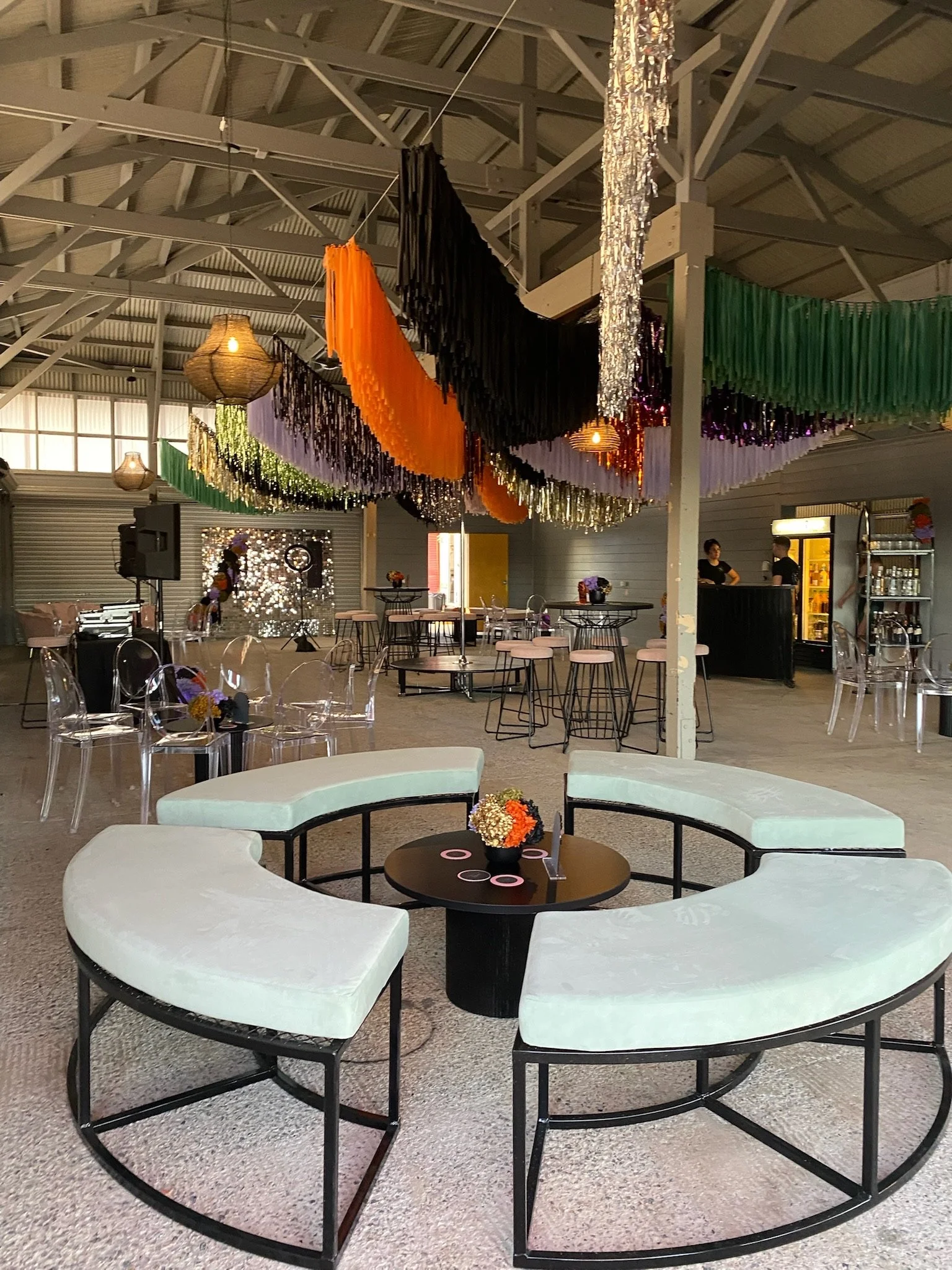 Decorated indoor party venue with colorful hanging streamers, tables, transparent chairs, and a seating area with curved ottomans around a round table with flowers.