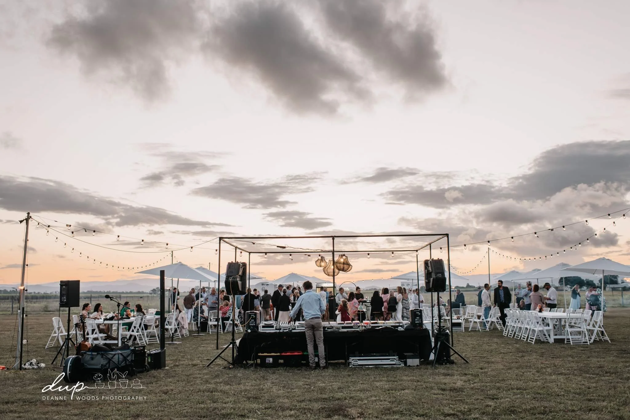 Outdoor wedding reception setup during sunset with guests, white tables and chairs, string lights, large umbrellas, and DJ equipment.