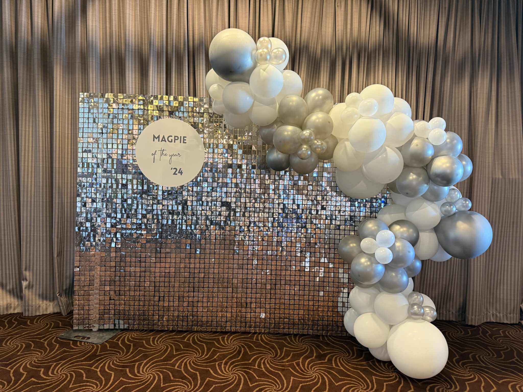 Decorative backdrop made of small reflective tiles with a cluster of white and silver balloons on the right side, and a circular sign with the text "MAGPIE of the year '24" in the center.