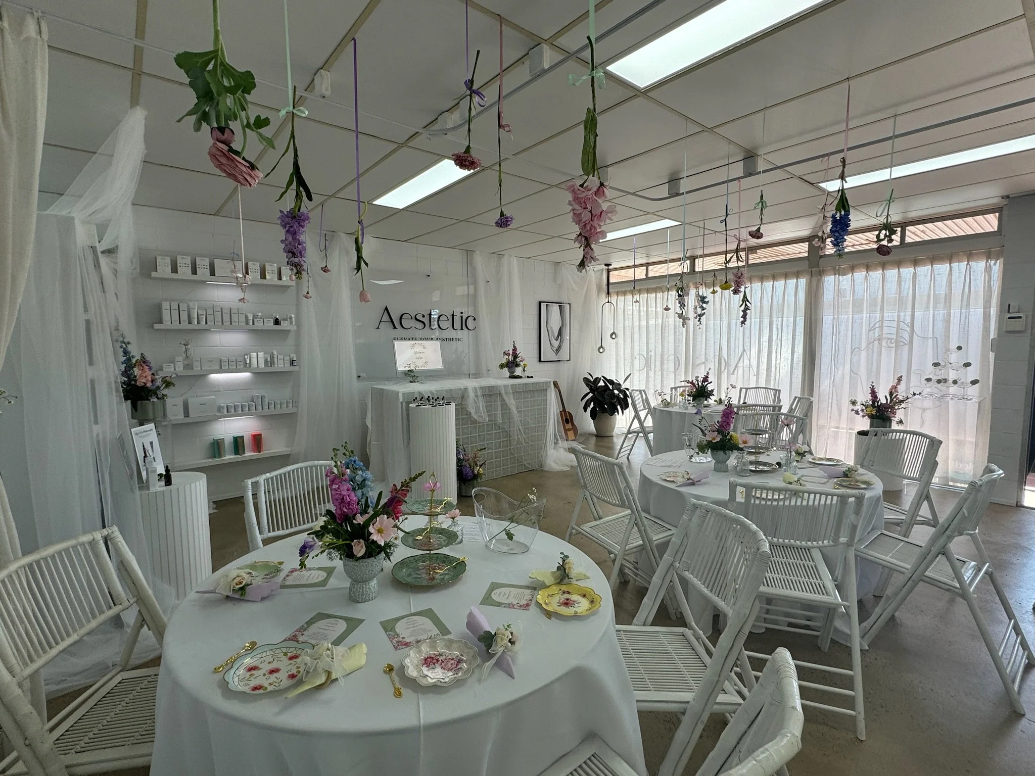 A beautifully decorated event space with white chairs and tables, floral centerpieces, hanging flowers from the ceiling, and white sheer curtains covering large windows.