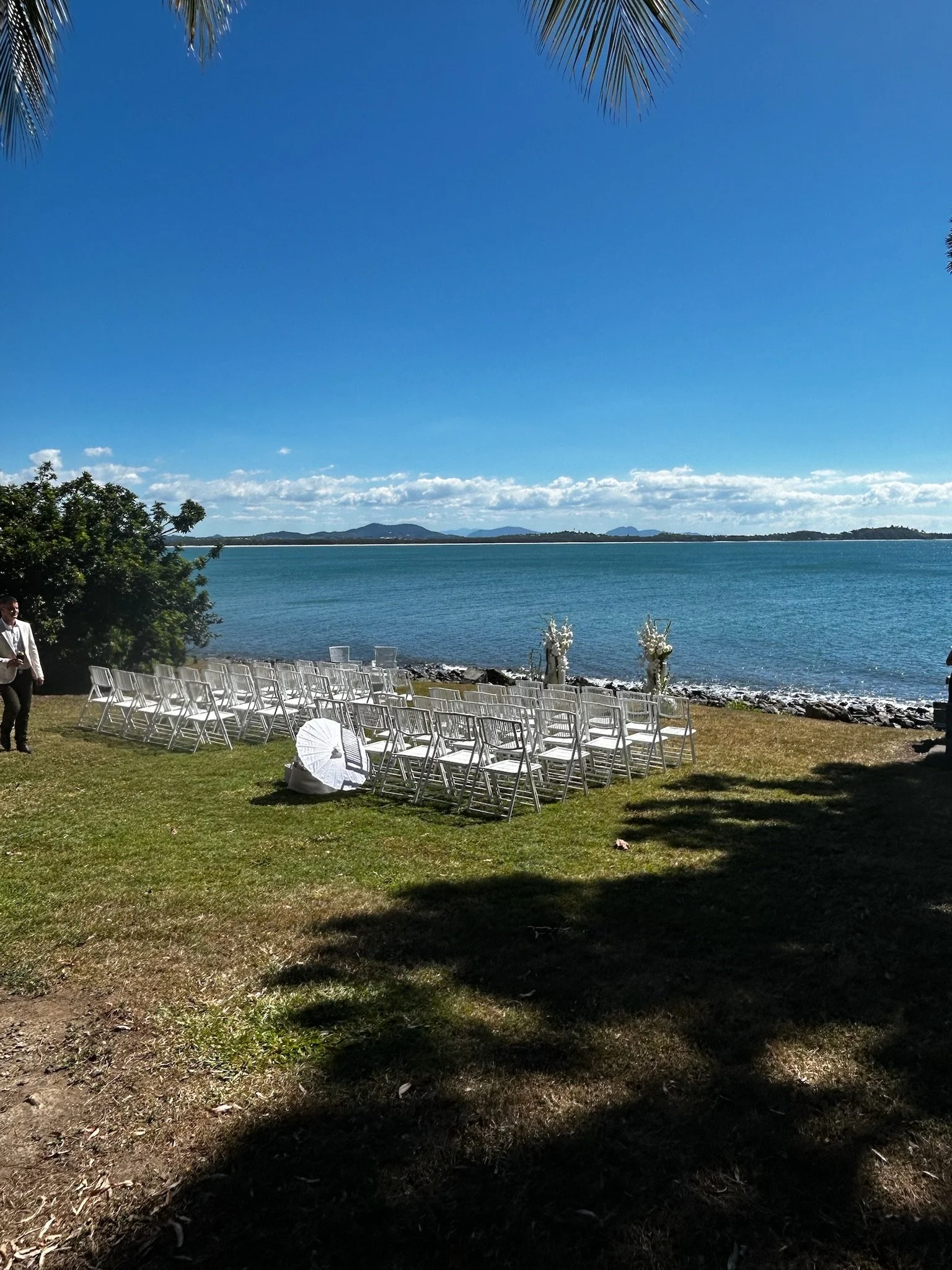 Outdoor wedding setup on a grassy area near a body of water, with rows of white chairs, flowers, and decorative elements, under a clear blue sky with some clouds and distant mountains in the background.