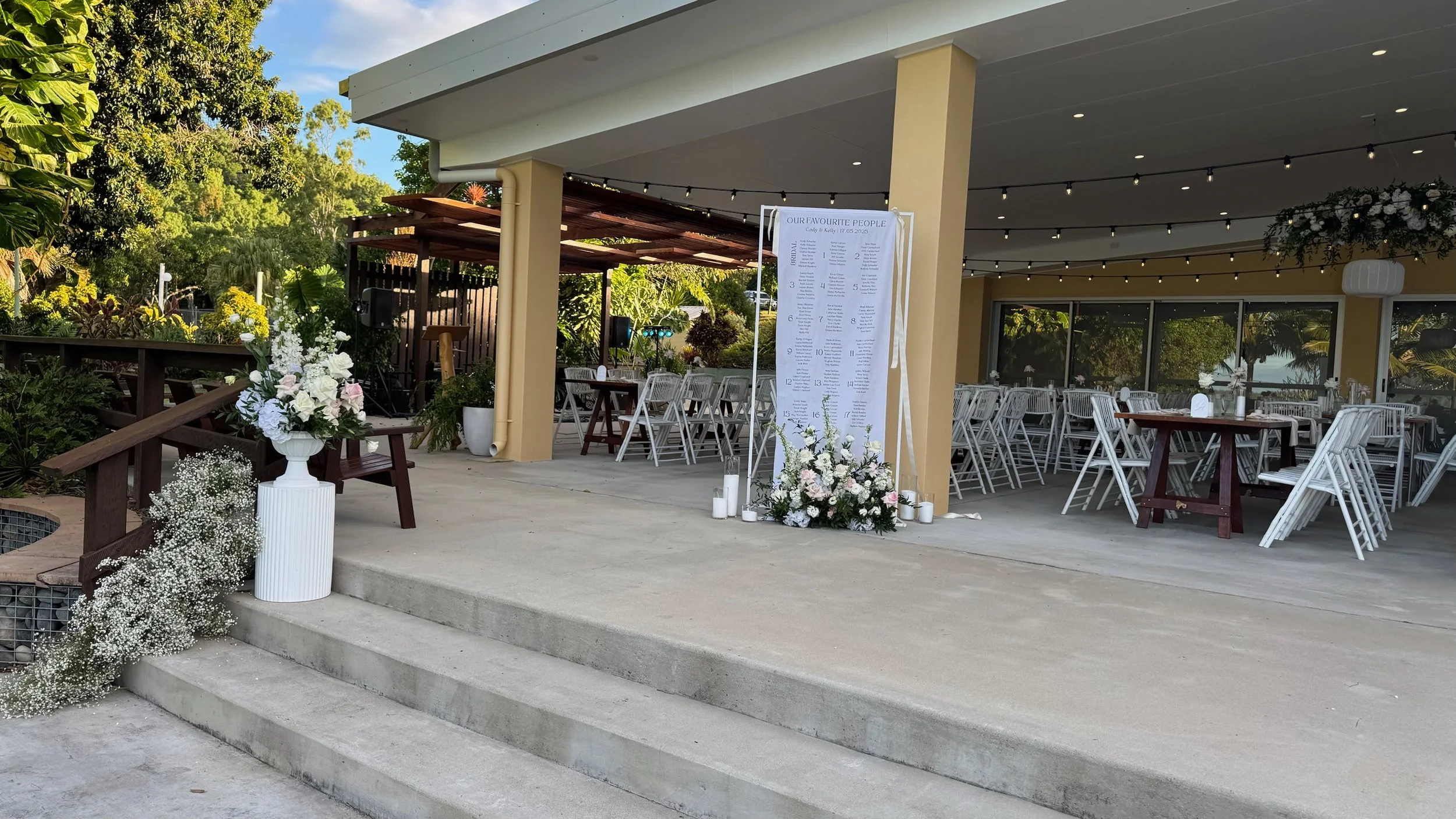 Outdoor event space decorated with white and pink flowers, tables, and chairs, set up under a covered patio with string lights, for a wedding or celebration.