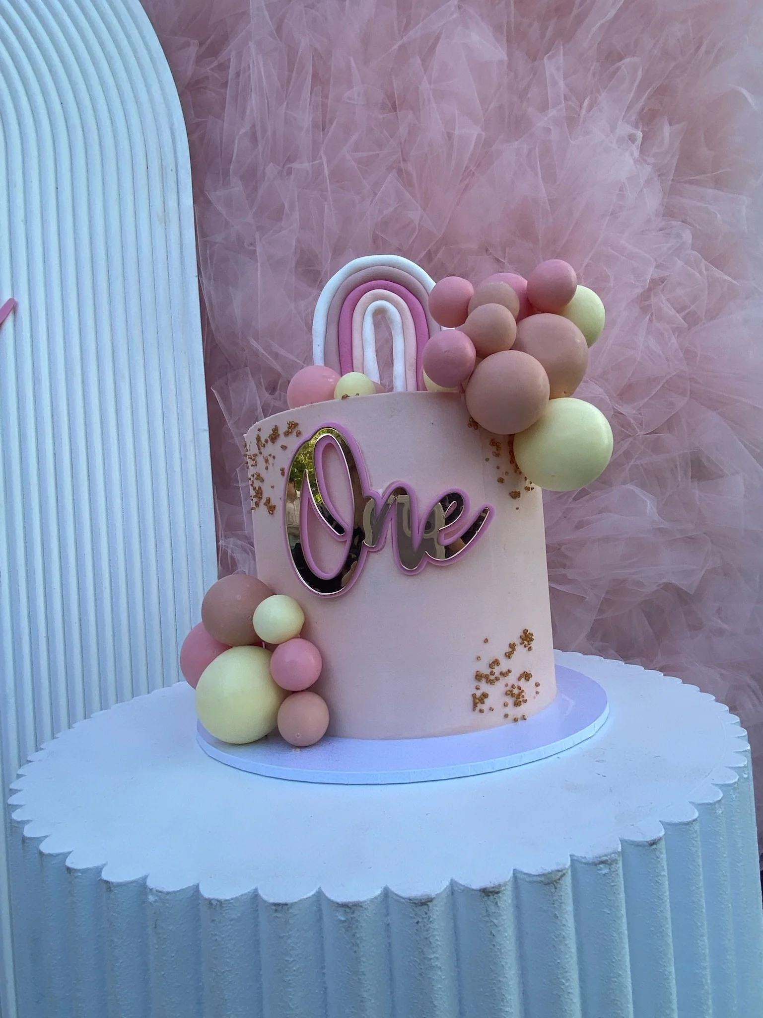 Pink birthday cake with pastel-colored decorations, including a rainbow topper, clusters of pink, yellow, and peach balls, gold sprinkles, and a silver and pink 'One' sign, on a white scalloped-edged cake stand against a pink tulle backdrop.