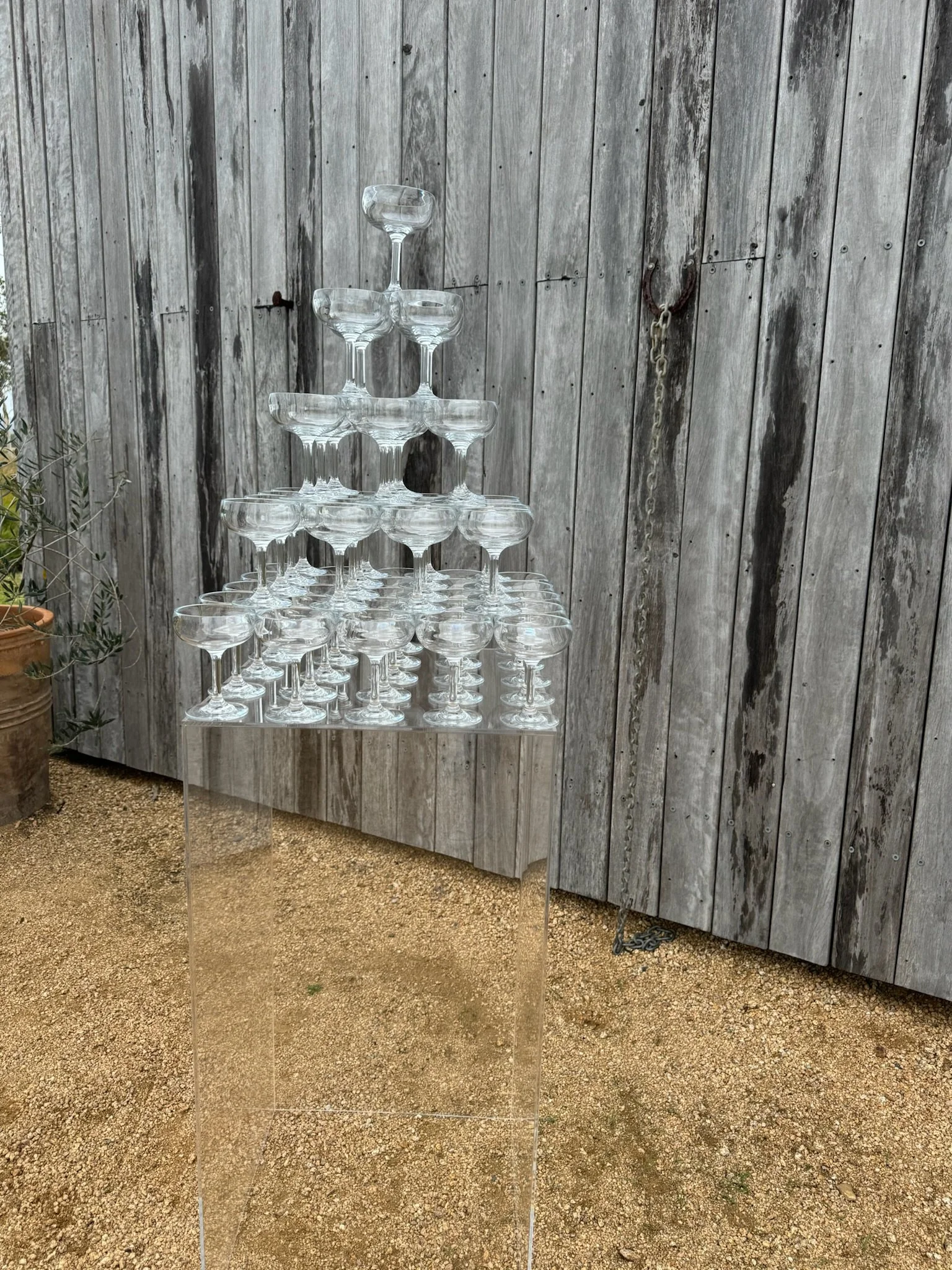 A pyramid of stacked empty champagne glasses arranged on a clear acrylic table outdoors with a weathered wooden fence in the background.
