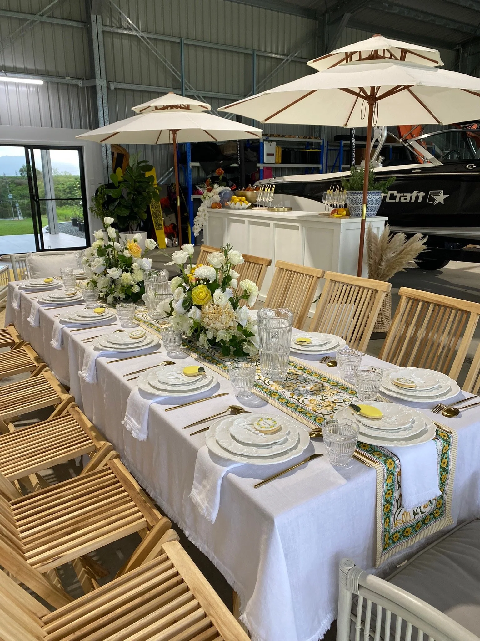 An indoor party setup featuring a long dining table with white tablecloth, decorated with floral centerpieces and place settings with plates, glasses, and gold utensils. Large cream-colored umbrellas provide shade over the table. In the background, t