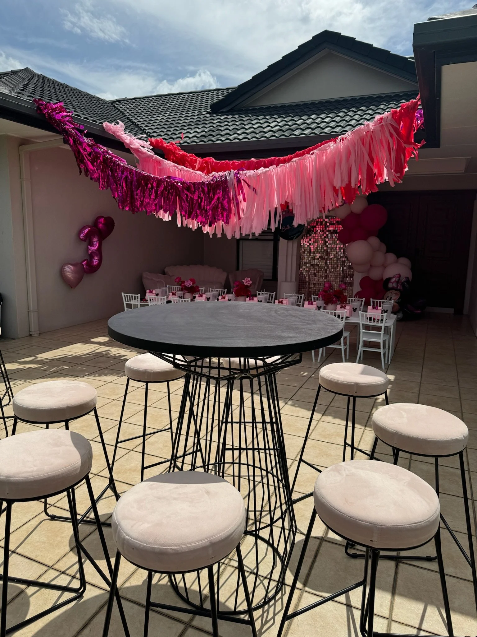 Decorated outdoor patio for a celebration with pink and red tassel banners hanging above, pink balloons on the wall, and a seating area with round tables and chairs, and a balloon arch in the background.