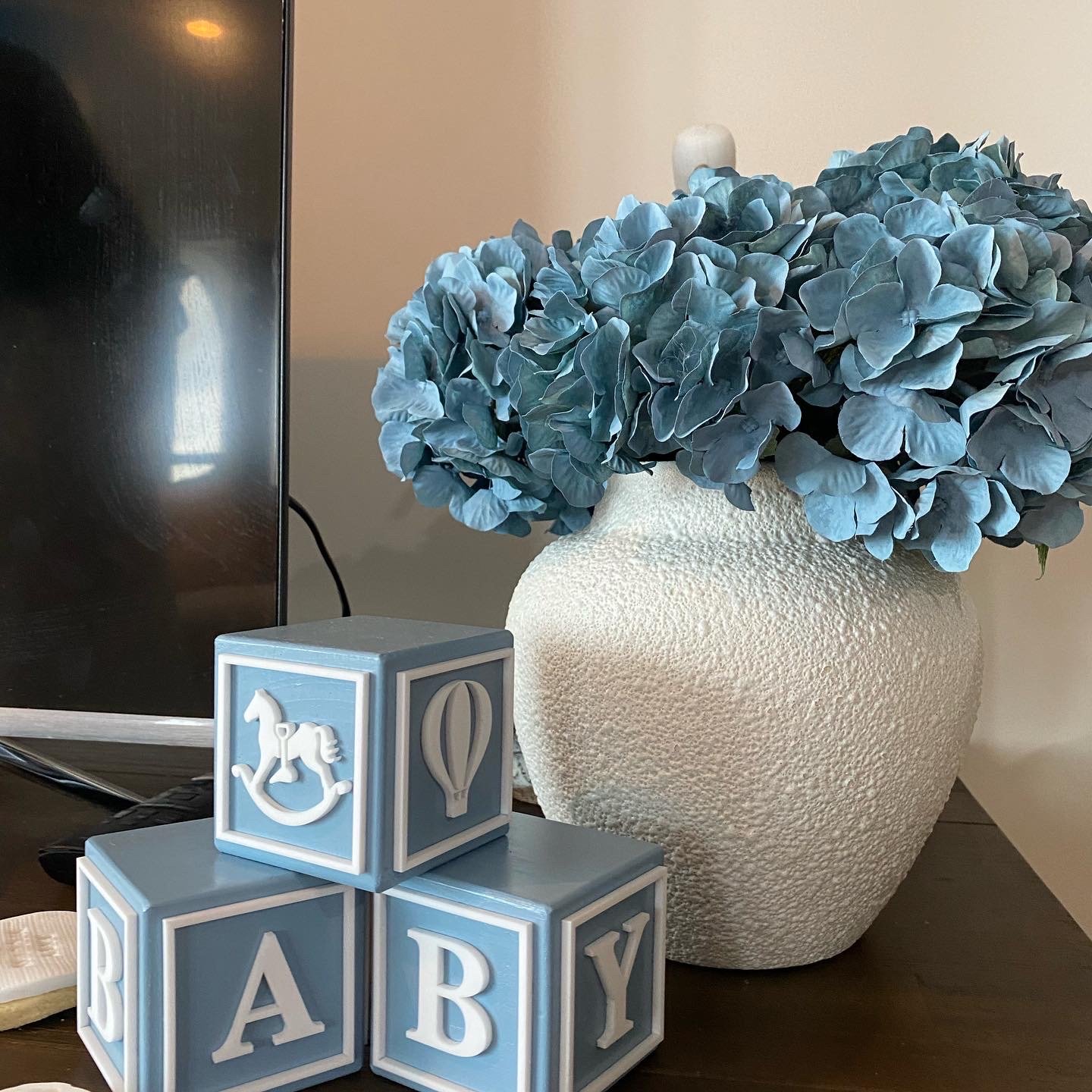 Blue hydrangea flowers in a textured white ceramic vase on a dark wooden surface, with blue and white baby blocks spelling 'BABY' in front of the vase, and a television screen visible on the left side.