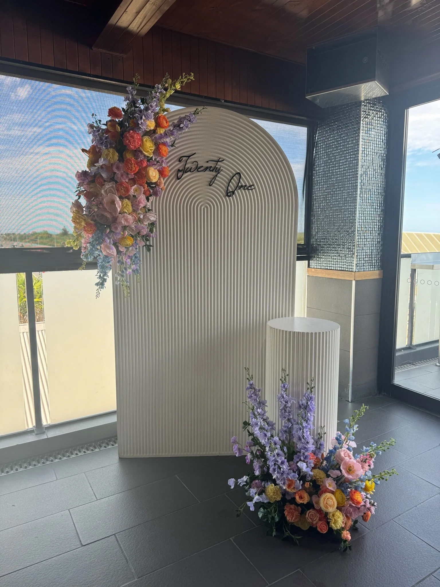 A floral display with pink, purple, yellow, and orange flowers arranged on a stand with the words 'Twenty One' written above, likely for a celebration.