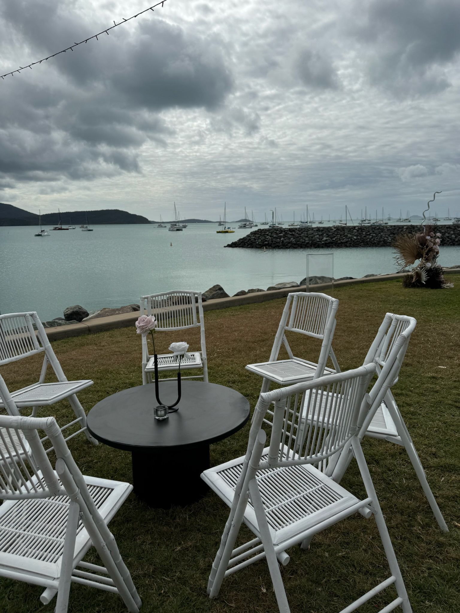 Outdoor seating area with white chairs around a black table by a body of water with boats and a cloudy sky.