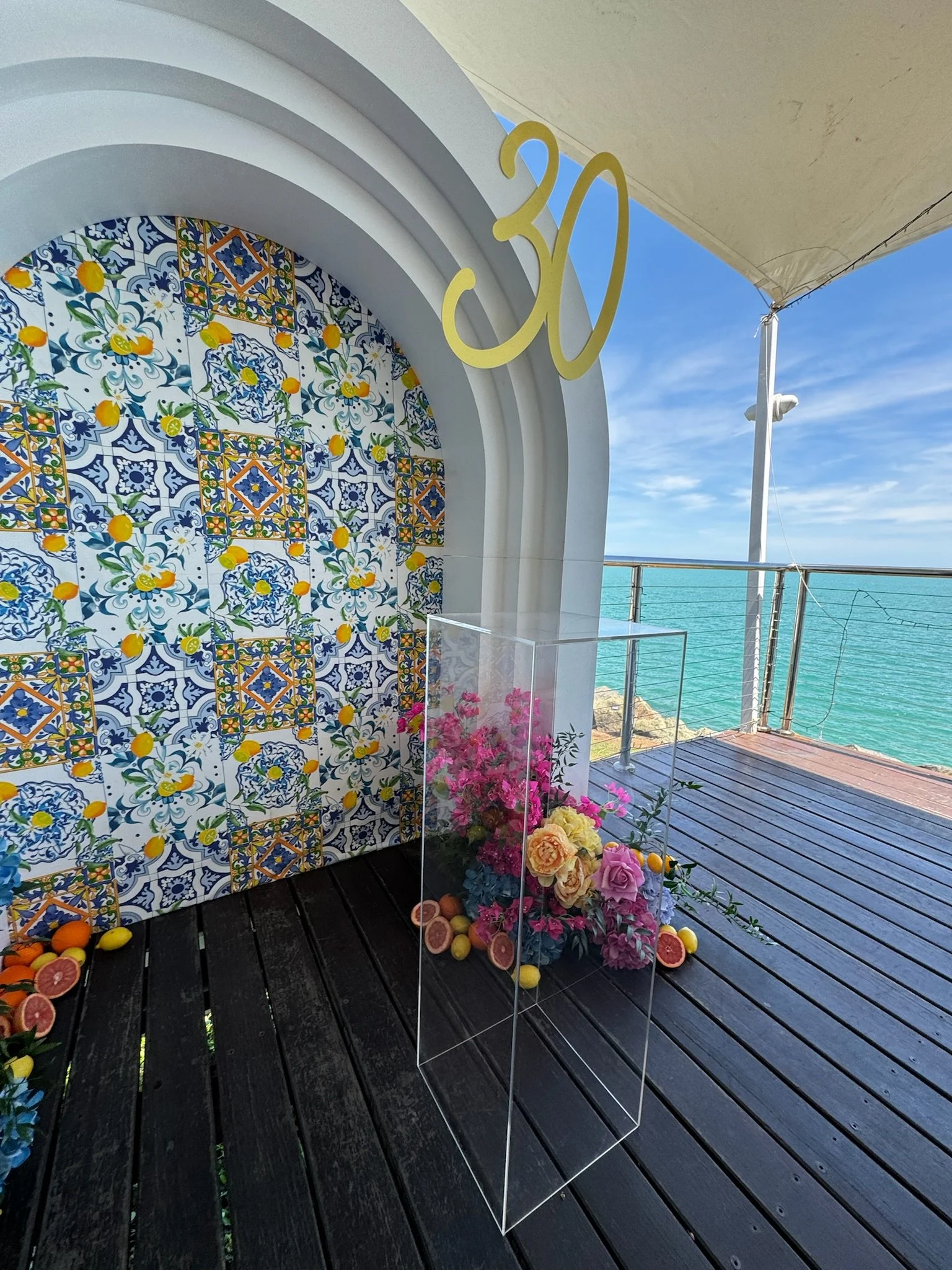 Decorative arch with a colorful patterned tile backdrop at an outdoor seaside location, featuring pink and yellow flowers in a glass display case, with scattered lemons and grapefruits on the dark wooden floor and a blue ocean view in the background,