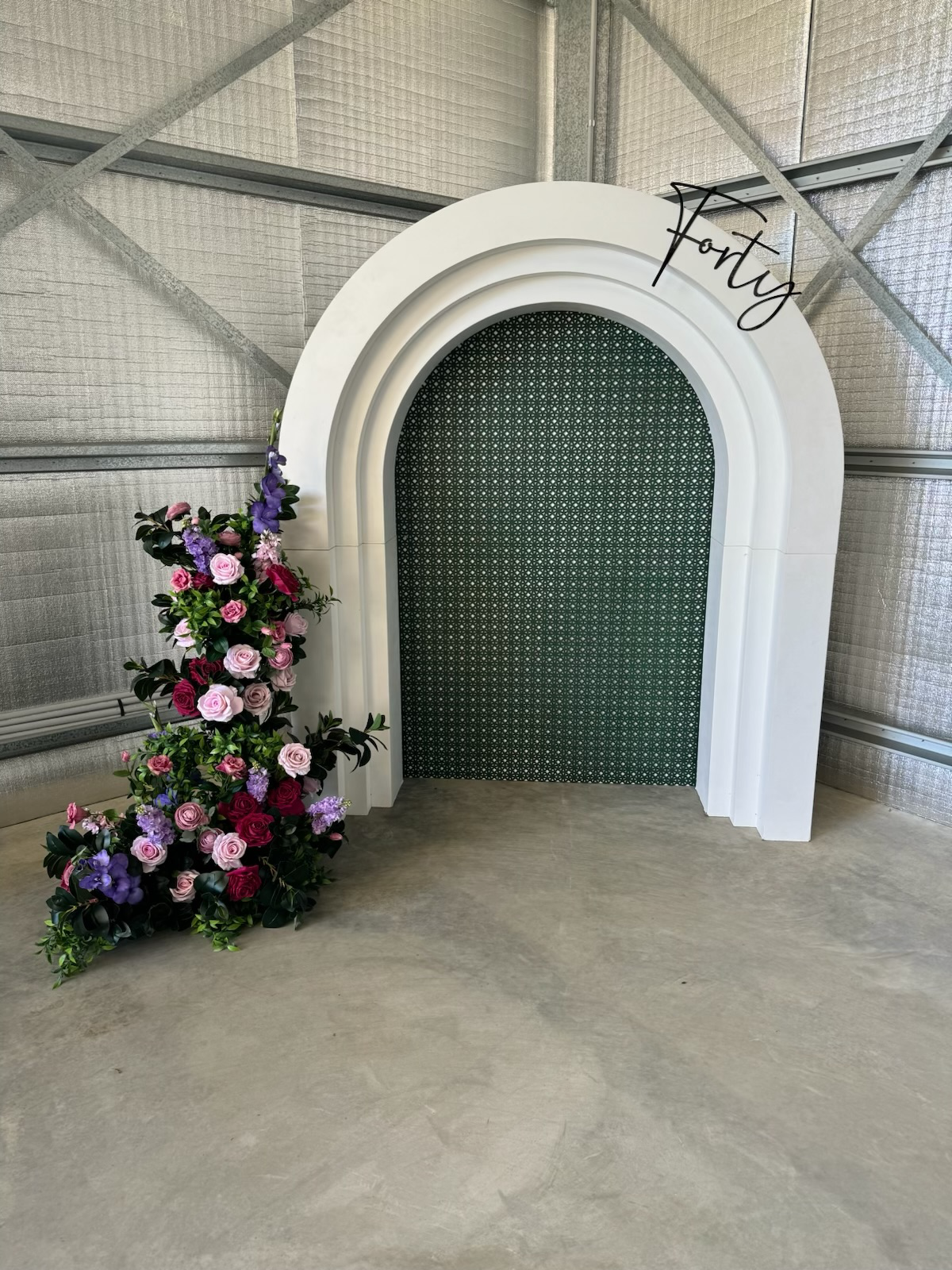 A decorative arch with greenery-patterned interior background, a floral arrangement with pink, purple, and red flowers on the side, and a sign that reads "Forty" in cursive on top.
