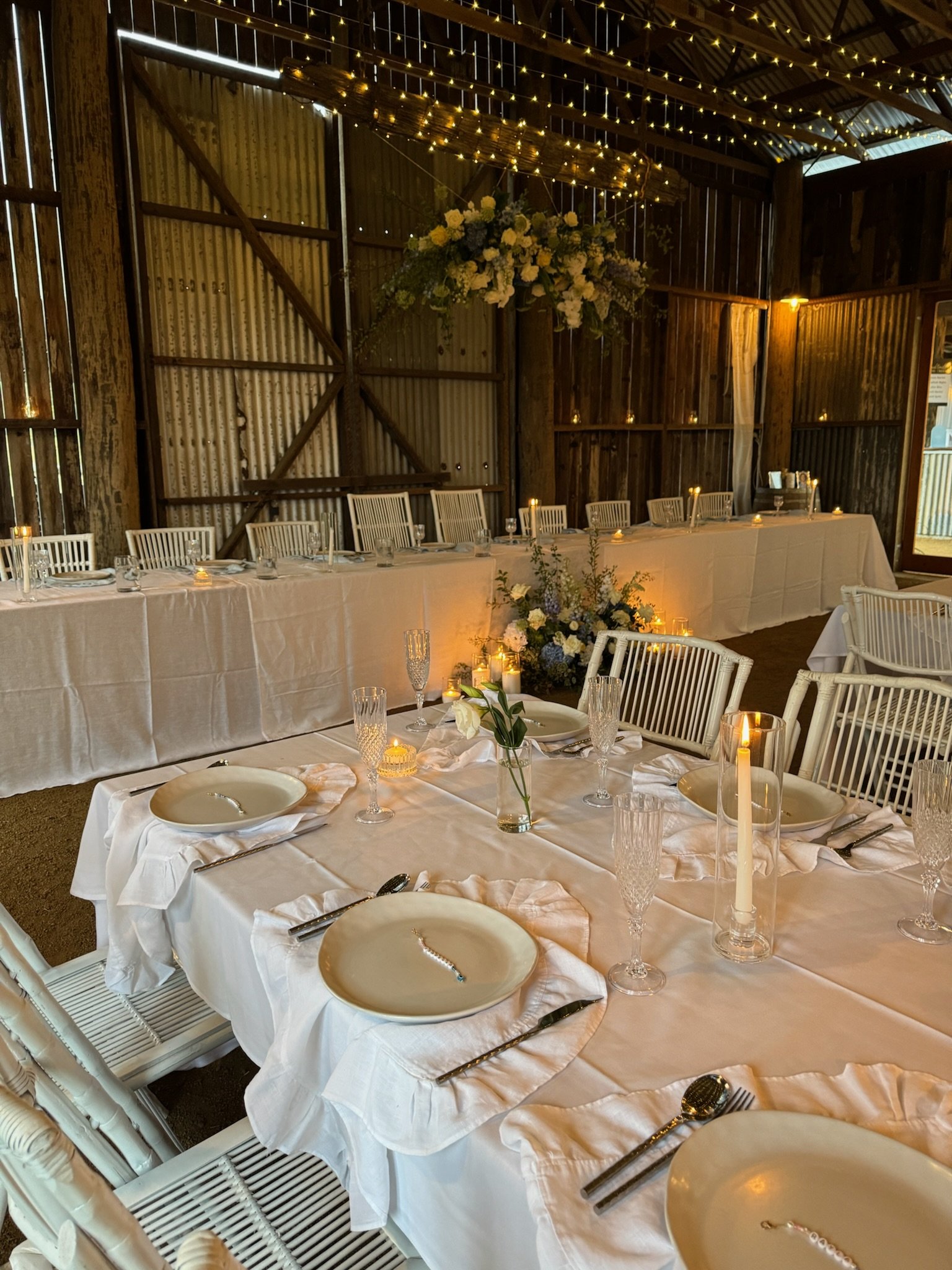 Elegant dining setup in a rustic barn with white tablecloths, brass candlesticks, floral centerpieces, and fairy lights.