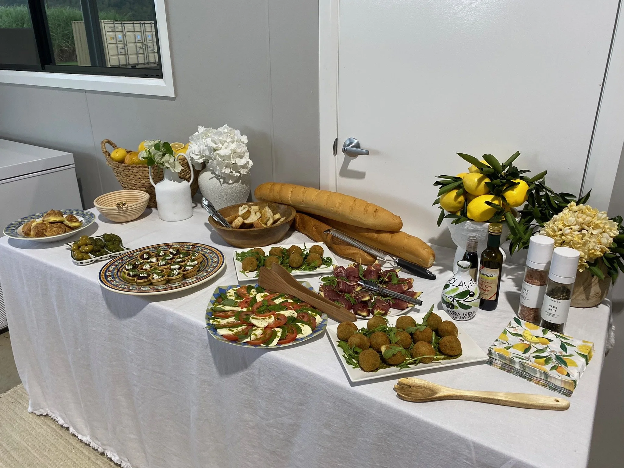 A table set with various foods including bread, cheeses, olives, tomatoes, prosciutto, and fried snacks, decorated with flowers, lemons, and bottles of oil and seasoning.