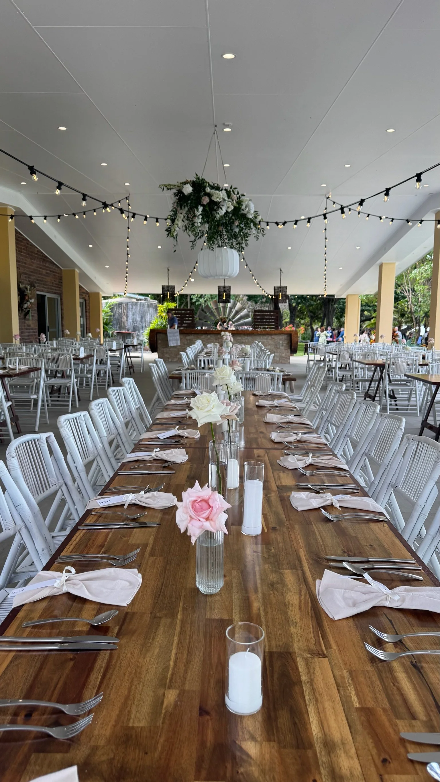 Long wooden table set for a wedding reception with white chairs, candles, and flowers. Overhead string lights and ceiling decorations with greenery and white flowers. In the background, a stage with DJ equipment and people outdoors.