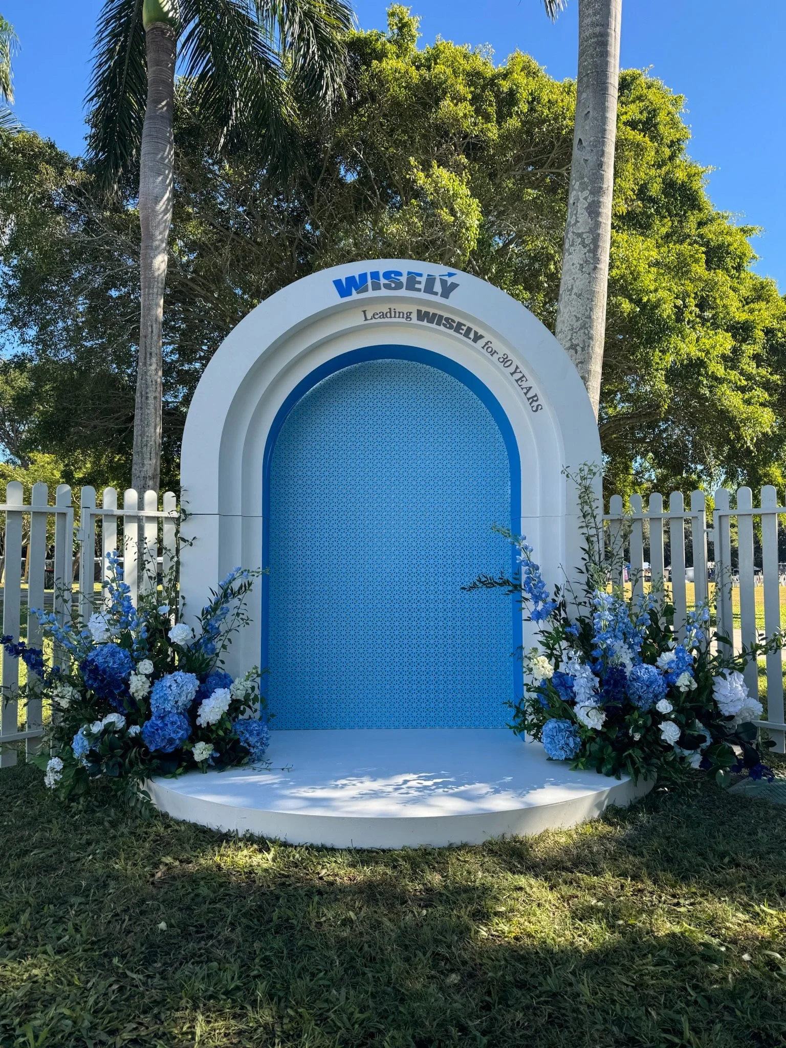 A decorative outdoor stage with blue floral arrangements on either side, white fencing in the background, and a large white and blue arch with the words 'WISELY' and 'Leading WISELY for 80 YEARS' written on it, set against a clear blue sky and green 