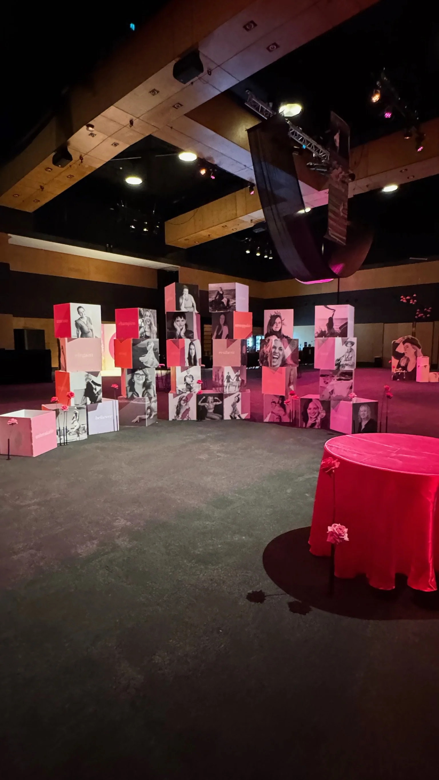 Event space with pink and white photo display cubes featuring images of women, a small round table with a red cloth, and pink flowers, under dim lighting.