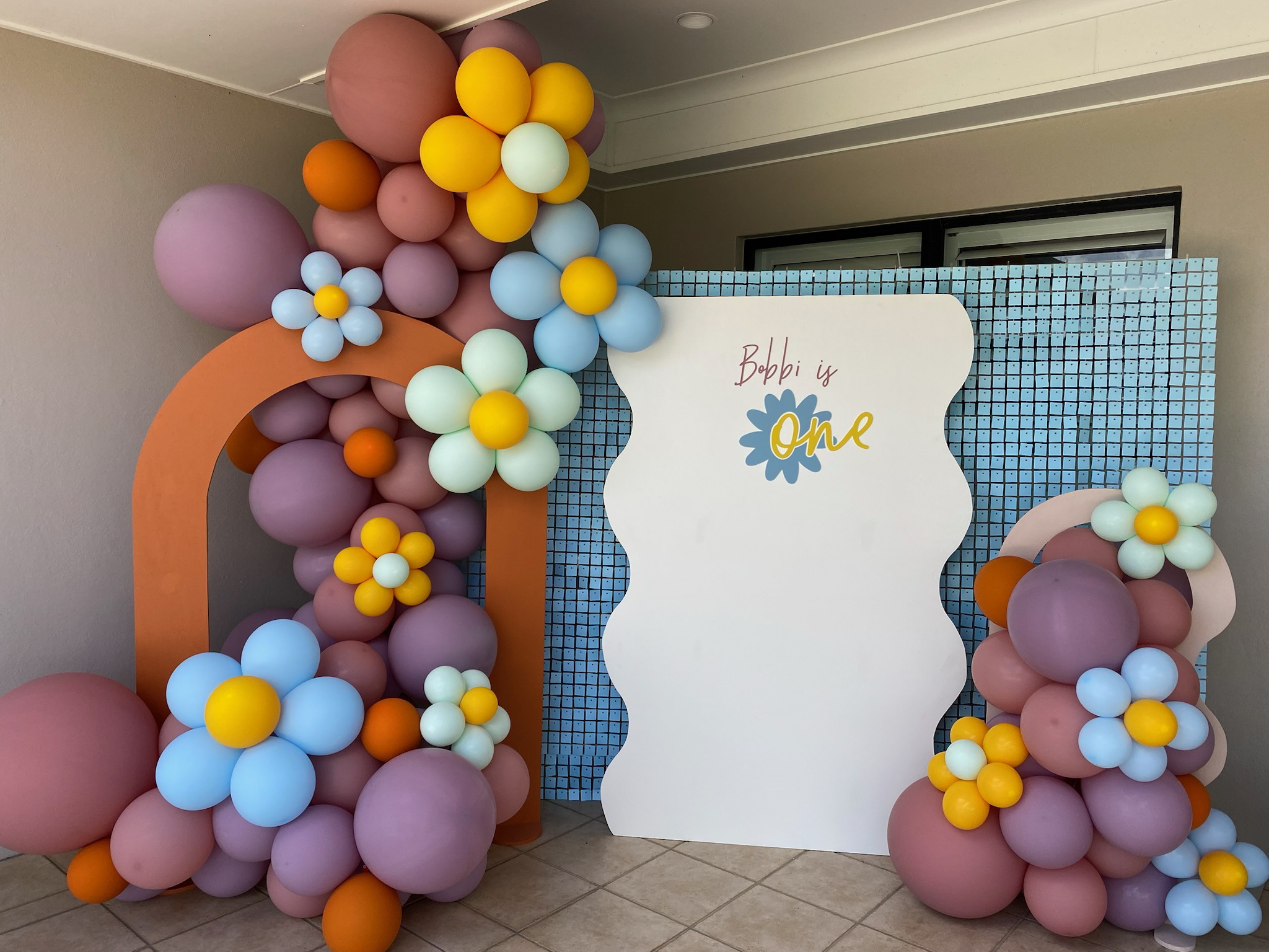 Decorations for a first birthday party with a balloon arrangement of pink, purple, yellow, orange, and white balloons, and a white board with the text "Bobbi is one" and a flower graphic, set against a blue grid backdrop.