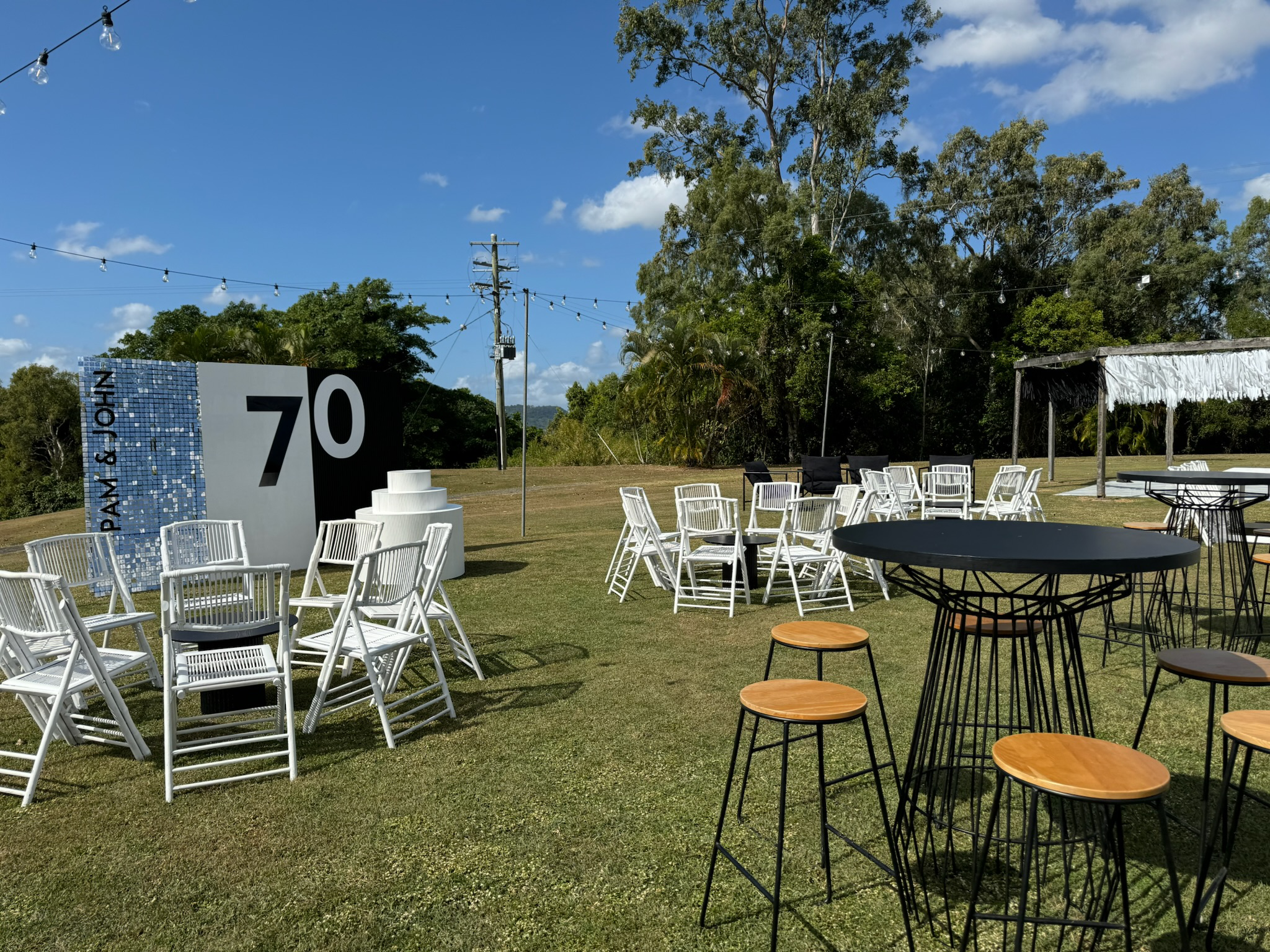 Outdoor event setup with white chairs, black high tables with wooden stools, and a large sign displaying the number 70 and "PINK GIN" on a blue mosaic-patterned background, under a clear blue sky with string lights and trees in the background.