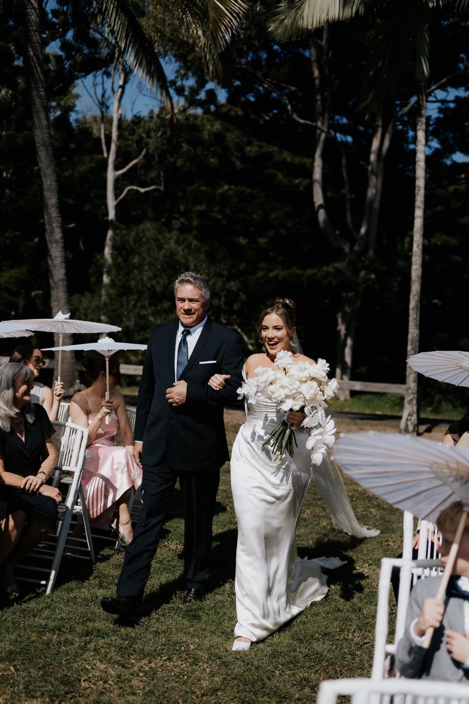 Bride walking down the aisle with her father at an outdoor wedding ceremony, holding a bouquet of white flowers, surrounded by guests holding white umbrellas.