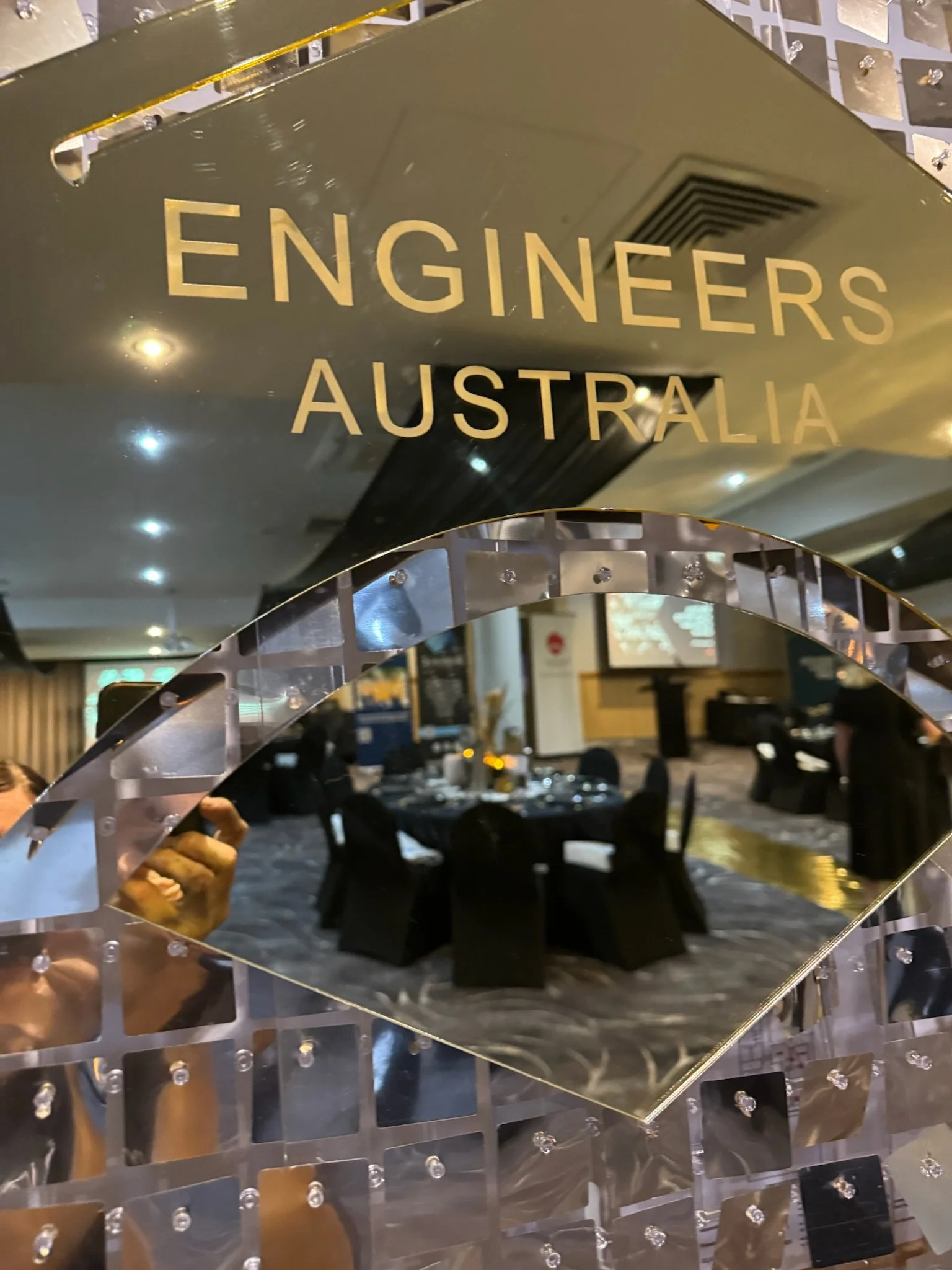 Mirror reflecting a banquet hall with round tables, black chairs, and gold accents, with a sign reading 'Engineers Australia' in the foreground.