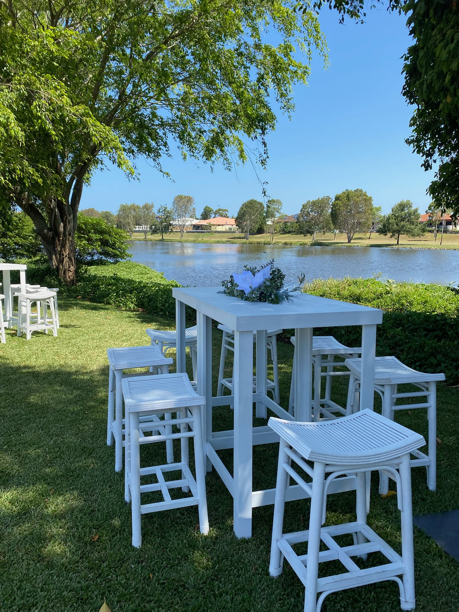 White outdoor bar table with white stools set up beside a lake, with trees and houses in the background, under a bright blue sky.